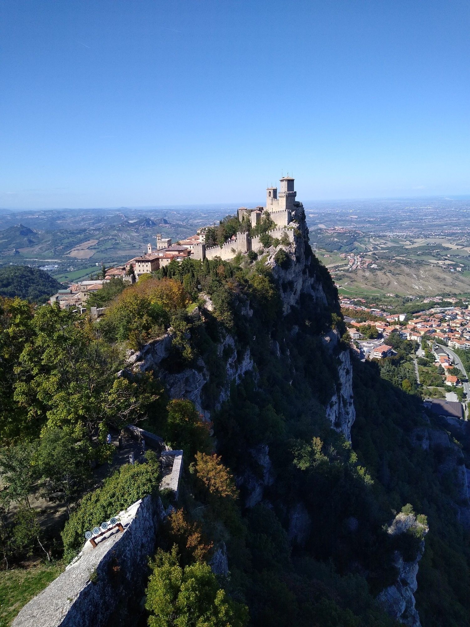 The first tower of San Marino, high atop Monte Titano. It is a clear bright day, and the medieval stone walls are built atop a sheer cliff with a village far down below. In the distance, the deep blue of the Adriatic Sea is barely visible