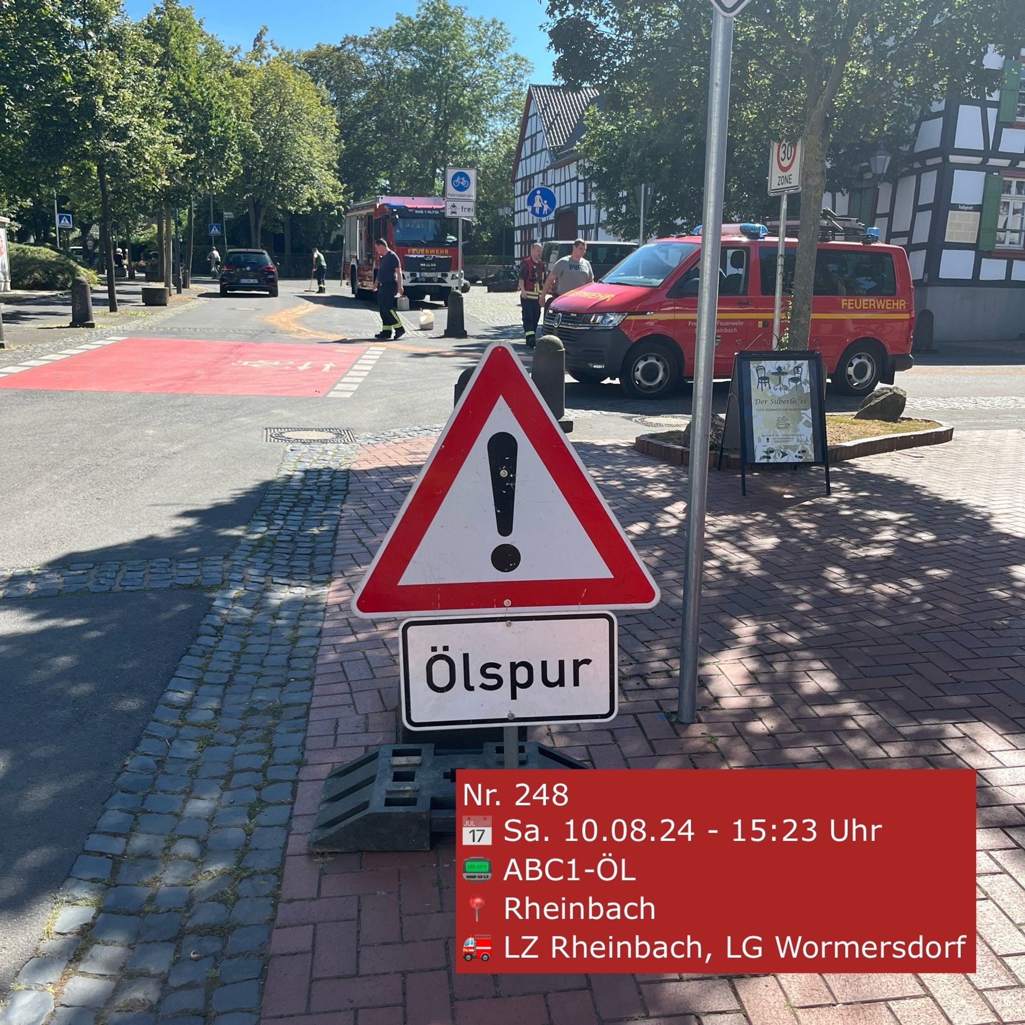 Ein Verkehrsschild "Gefahrstelle" und darunter der Hinweis "Ölspur" steht auf einer Straße. Im Hintergrund Einsatzfahrzeuge der Feuerwehr.