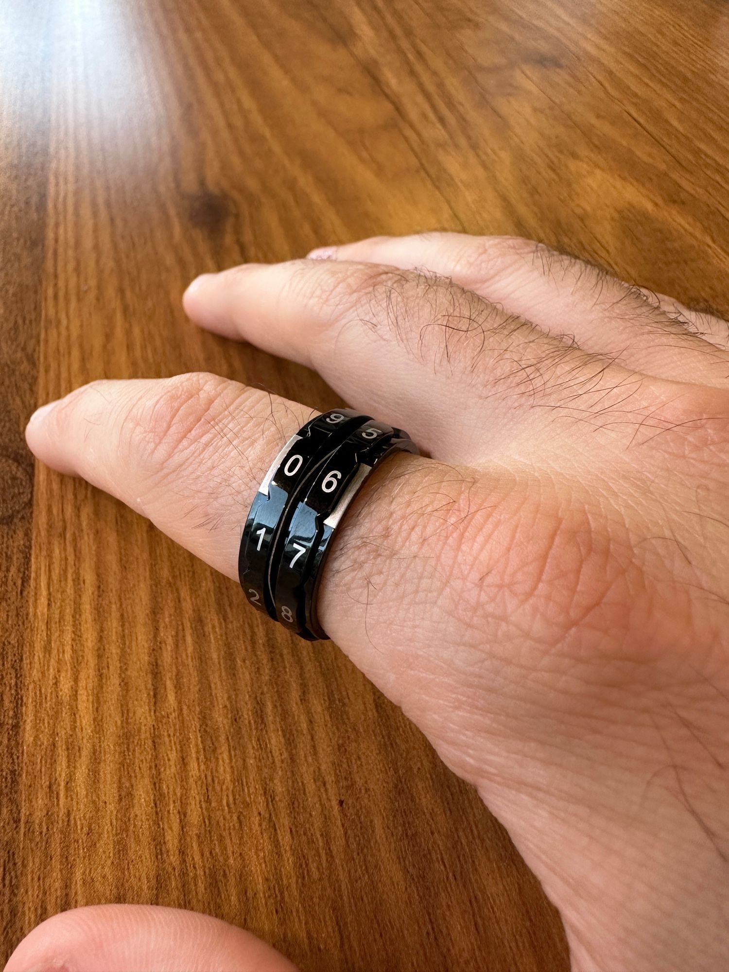 A hand resting on a wooden table. A ring on the index finger features two loops covered in numbers from 0 to 9. 