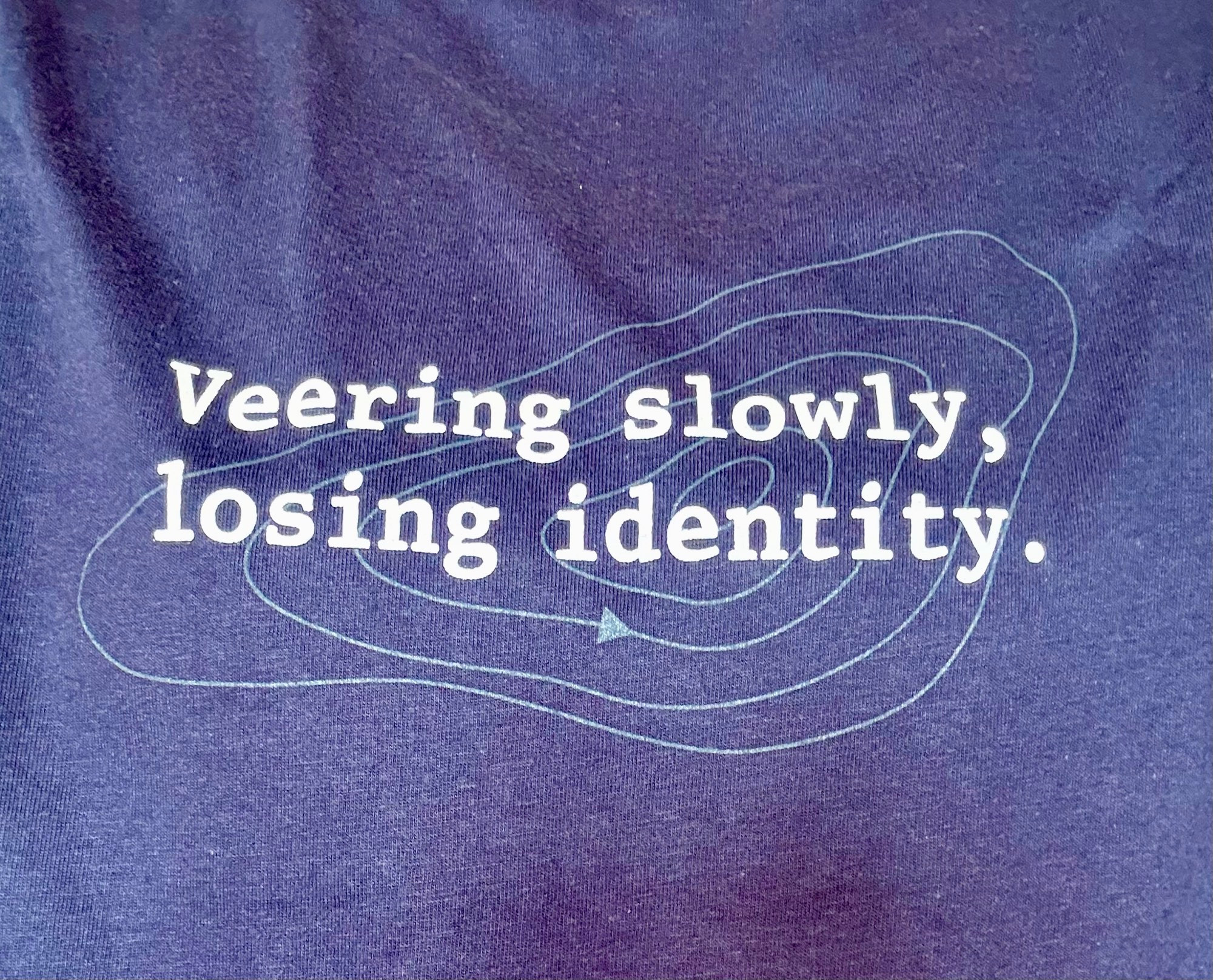 Close up on a navy t-shirt with a design of grey isobars and the words veering slowly, losing identity in white typewriter font 