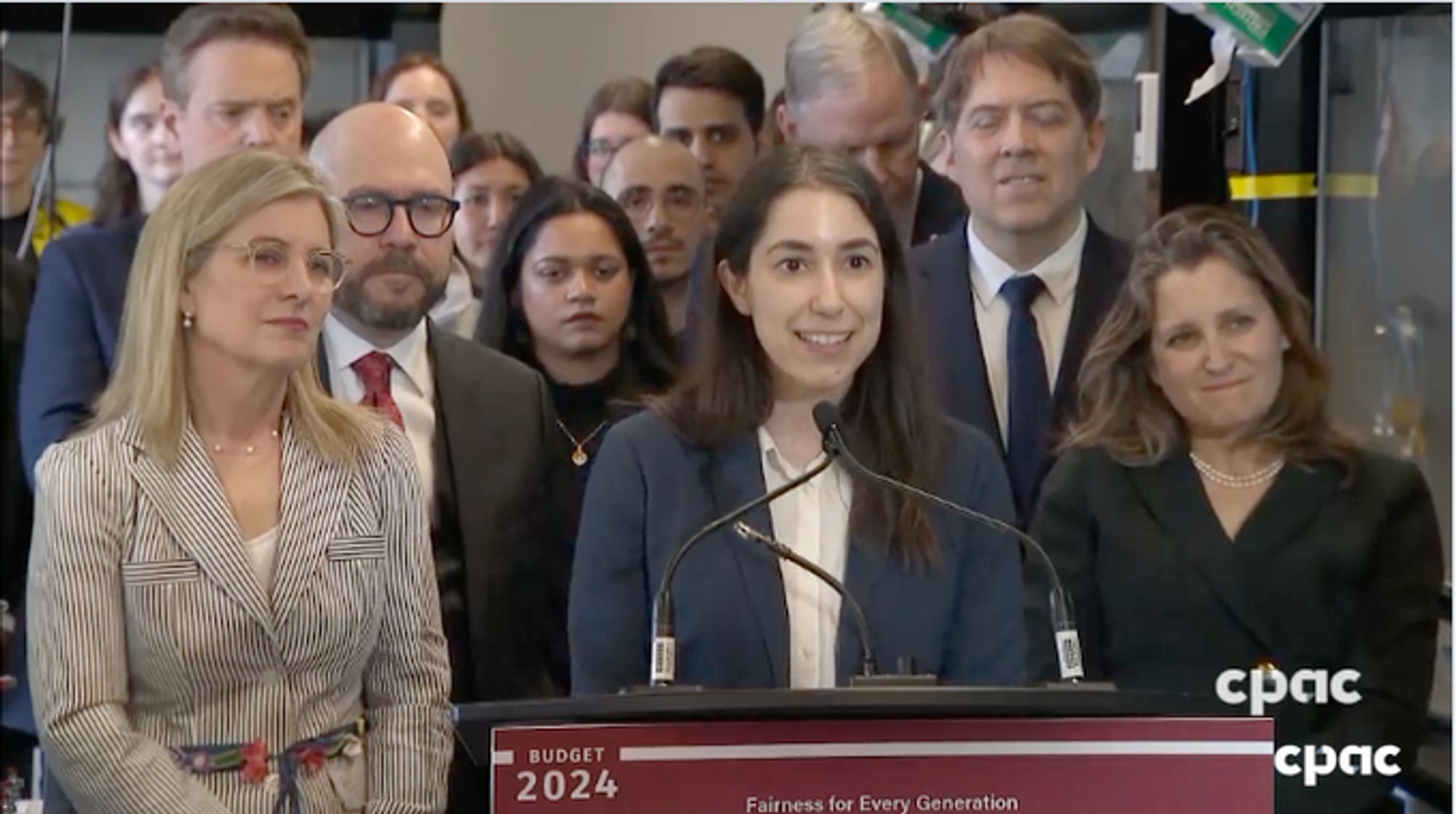 Women in business attire speaks at a lectern in a press conference surrounded by other people. To the right is Deputy Prime Minister Chrystia Freeland.