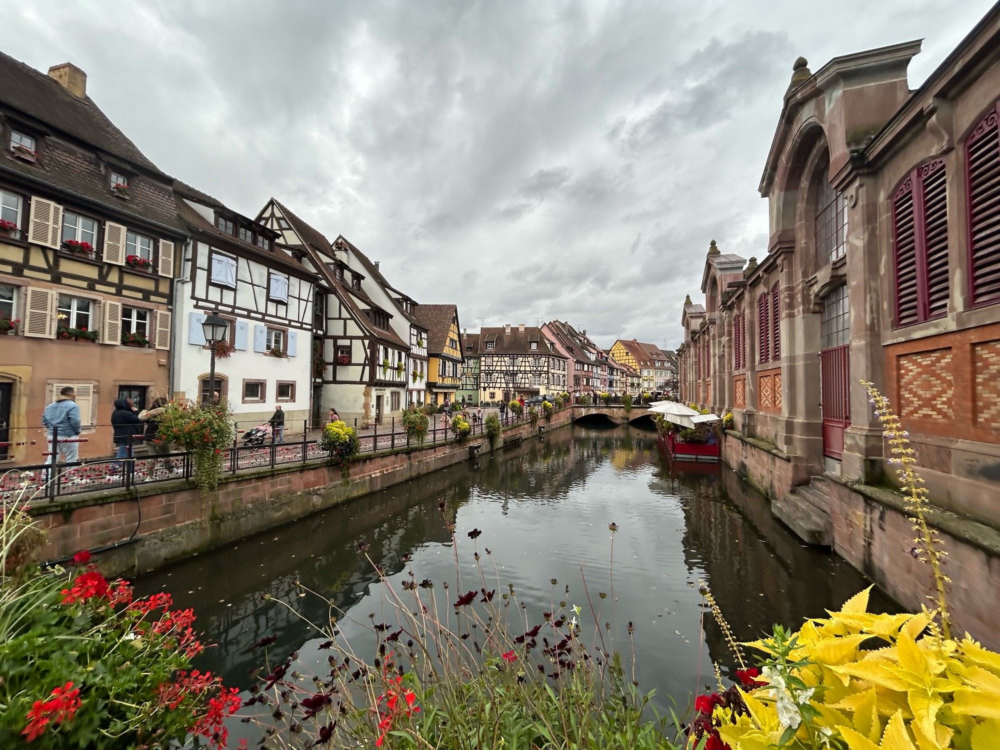 Blick von der von Blumen gesäumten Brücke einer Art Gracht auf eine Reihe Fachwerkhäuser.