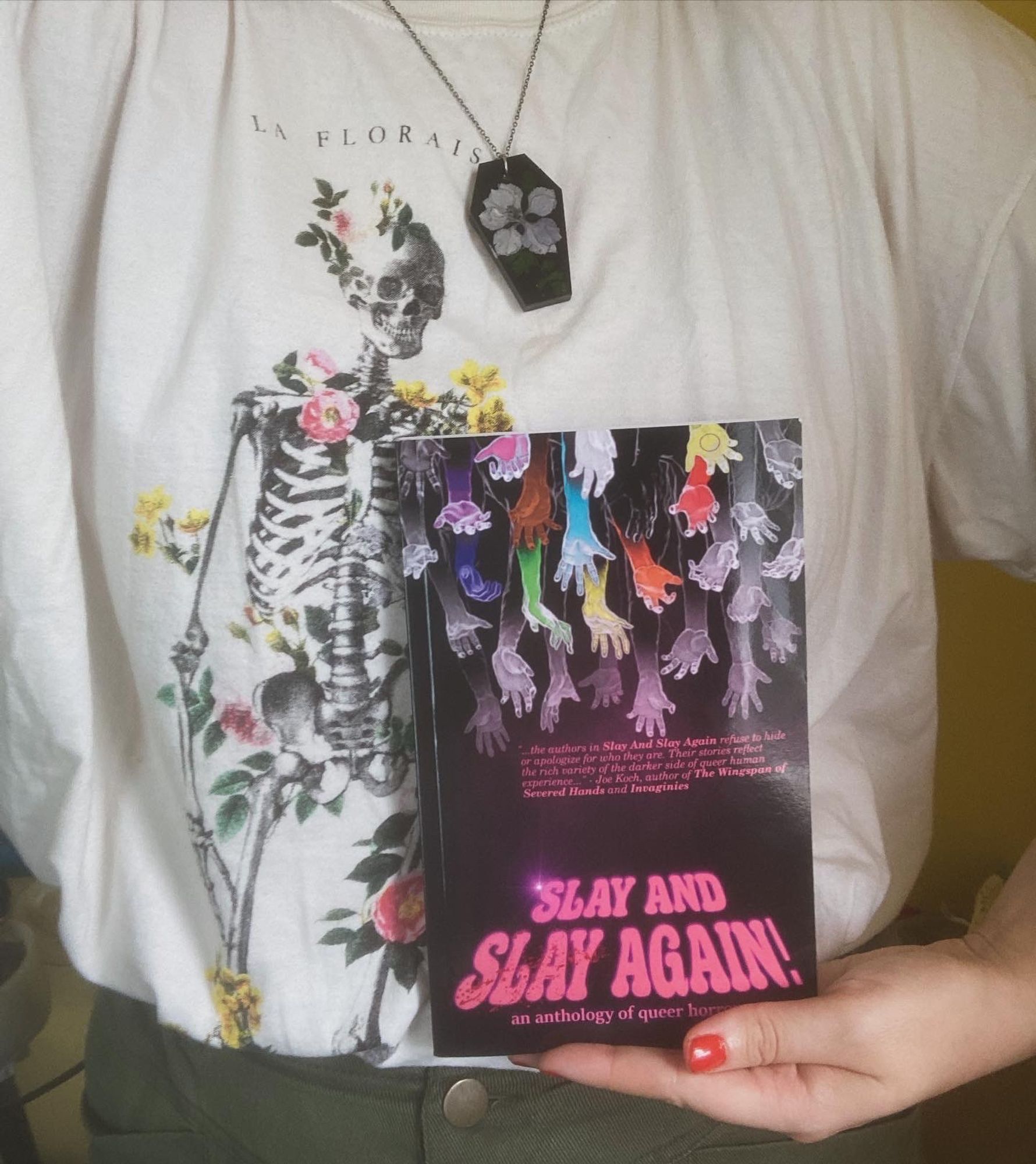 Photo of Marisca holding SLAY AND SLAY AGAIN. Visible: neck down to belt, wearing an oversized white tee with a graphic of a monochrome skeleton sprouting multicolored flowers. Marisca also wears a black resin coffin necklace with a purple flower. Cover art for SLAY AND SLAY AGAIN features a dark magenta background with rainbow zombie arms grasping down from the top towards the title in bold pink text.