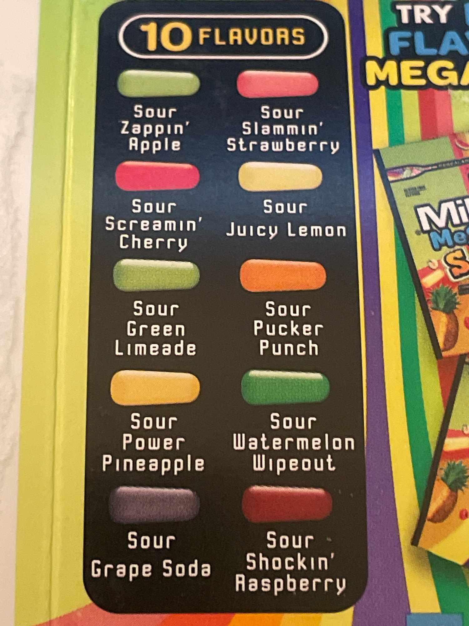 The packaging of sour Mike and Ikes candy, showing the list of flavors. green Sour Zappin' Apple, pink Sour Slammin' Strawberry, red Sour Screamin' Cherry, yellow Sour Juicy Lemon, green Sour Green Limeade, pink Sour Pucker Punch, yellow Sour Power Pineapple, green Sour Watermelon, purple Wipeout Sour Grape Soda, and dark red Sour Shockin' Raspberry.