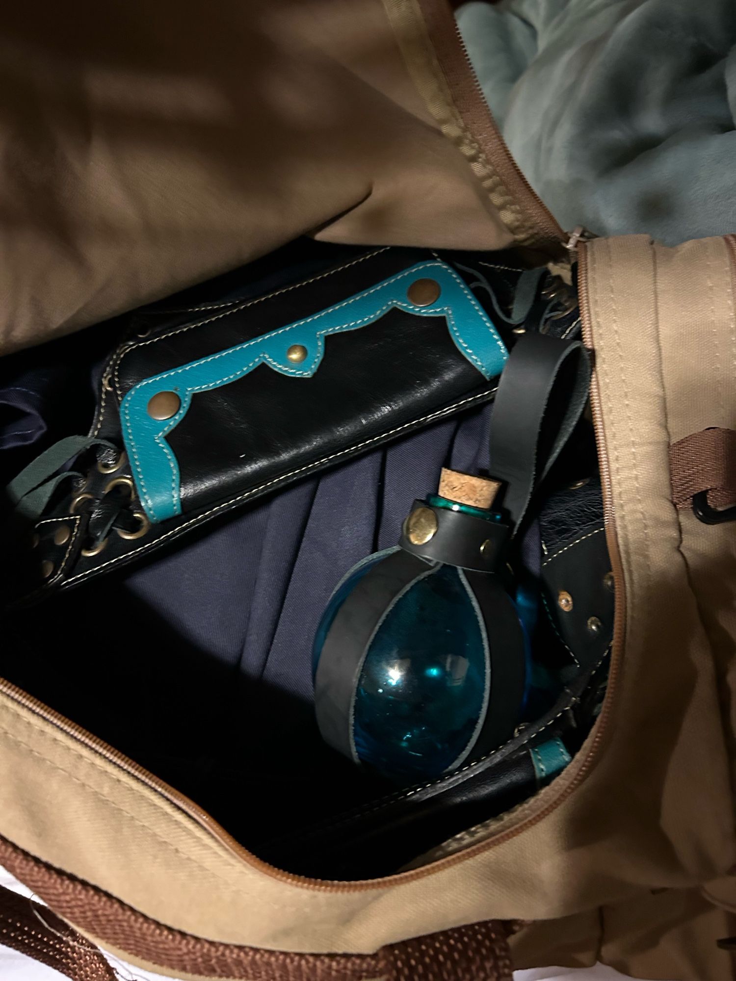 An open carrying bag with a folded leather belt laying over some ren fest garb that’s been tucked away.  On top is a blue glass bottle with a cork.