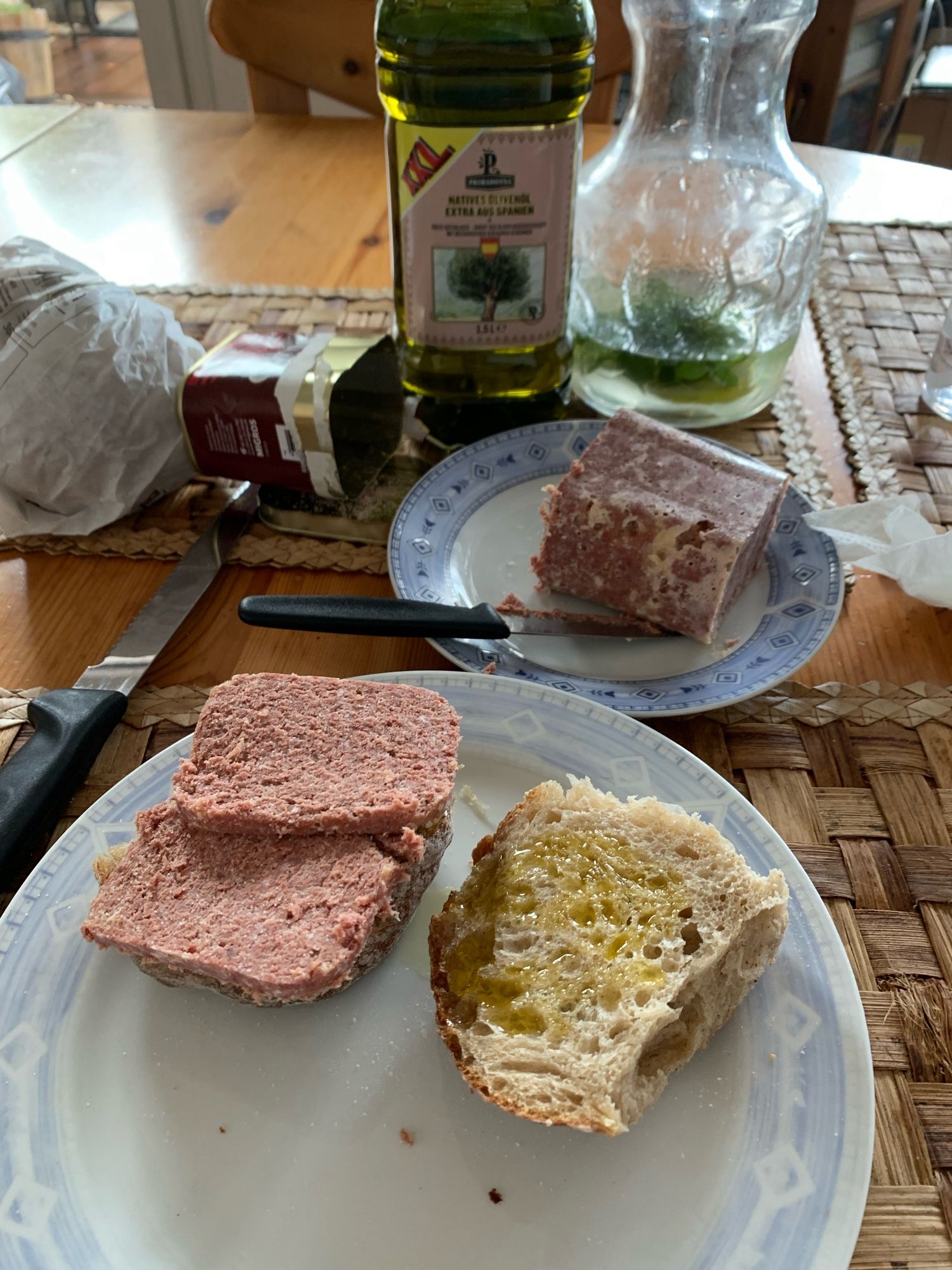 A picture of corn beef on bread and more bread with olive oil. In the center: corn beef on a plate. In the background is a bottle of olive oil and an empty can of corn beef next to a jug of water.