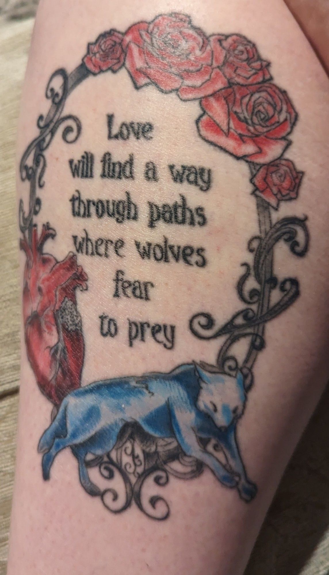 A tattoo made up of the words "Love will find a way through paths where wolves fear to prey" in an oval frame. At the top of the frame are red roses, to the left is an anatomical heart and at the bottom is a blue grey wolf