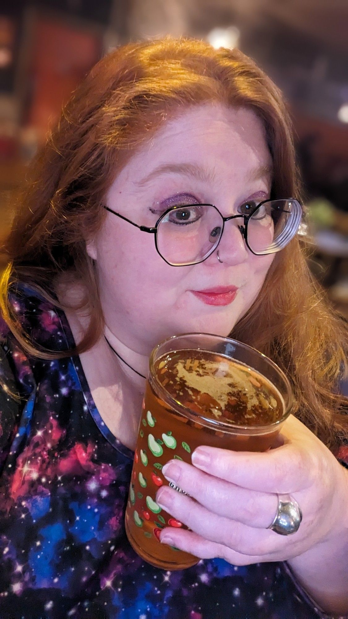 A red haired woman wearing a dress that looks like a bisexual galaxy is holding up a pint of cider