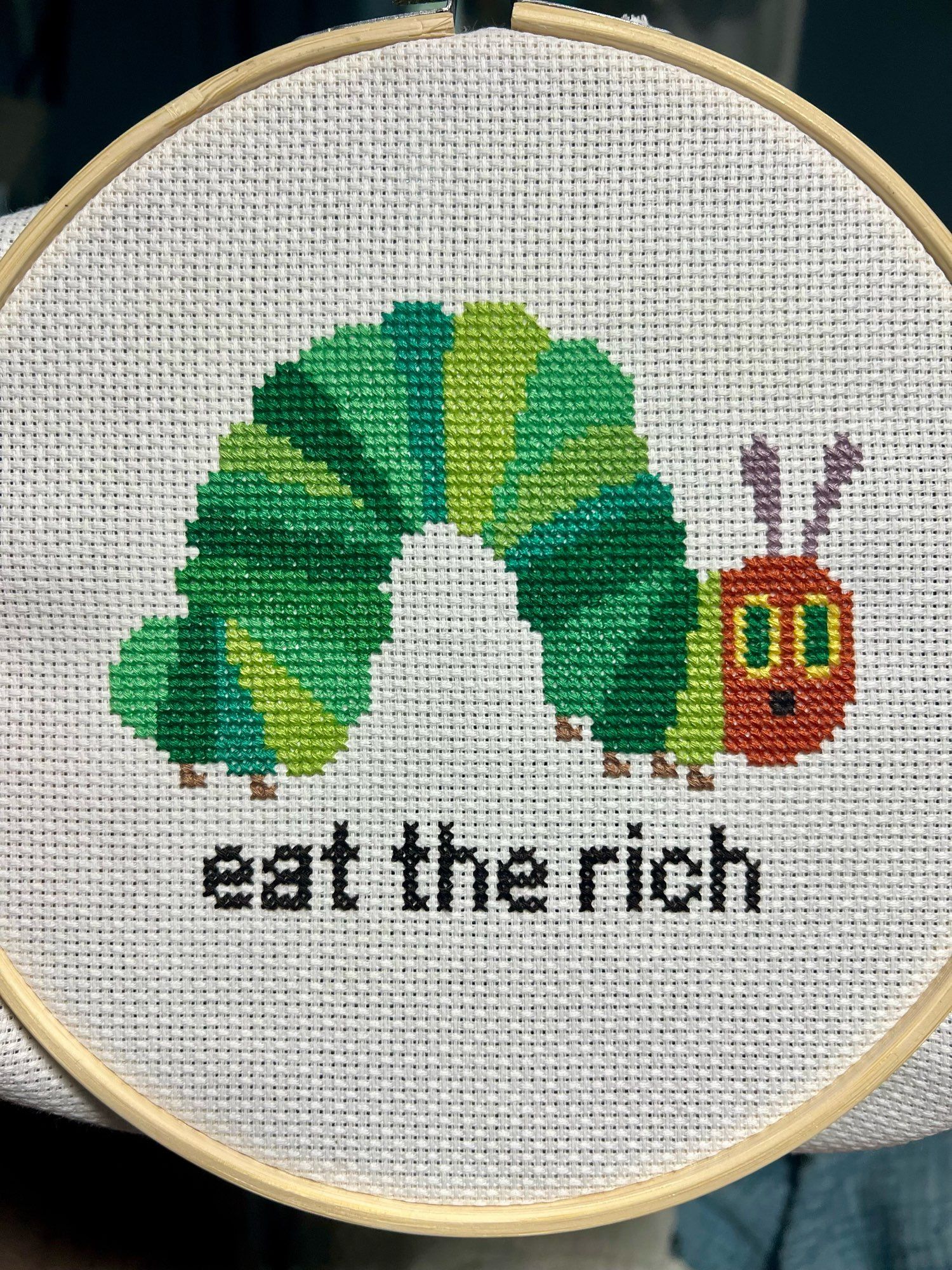 a cross-stitched version of The Very Hungry Caterpillar from Eric Carle’s classic children’s book of the same name from 1969. it is captioned “eat the rich”