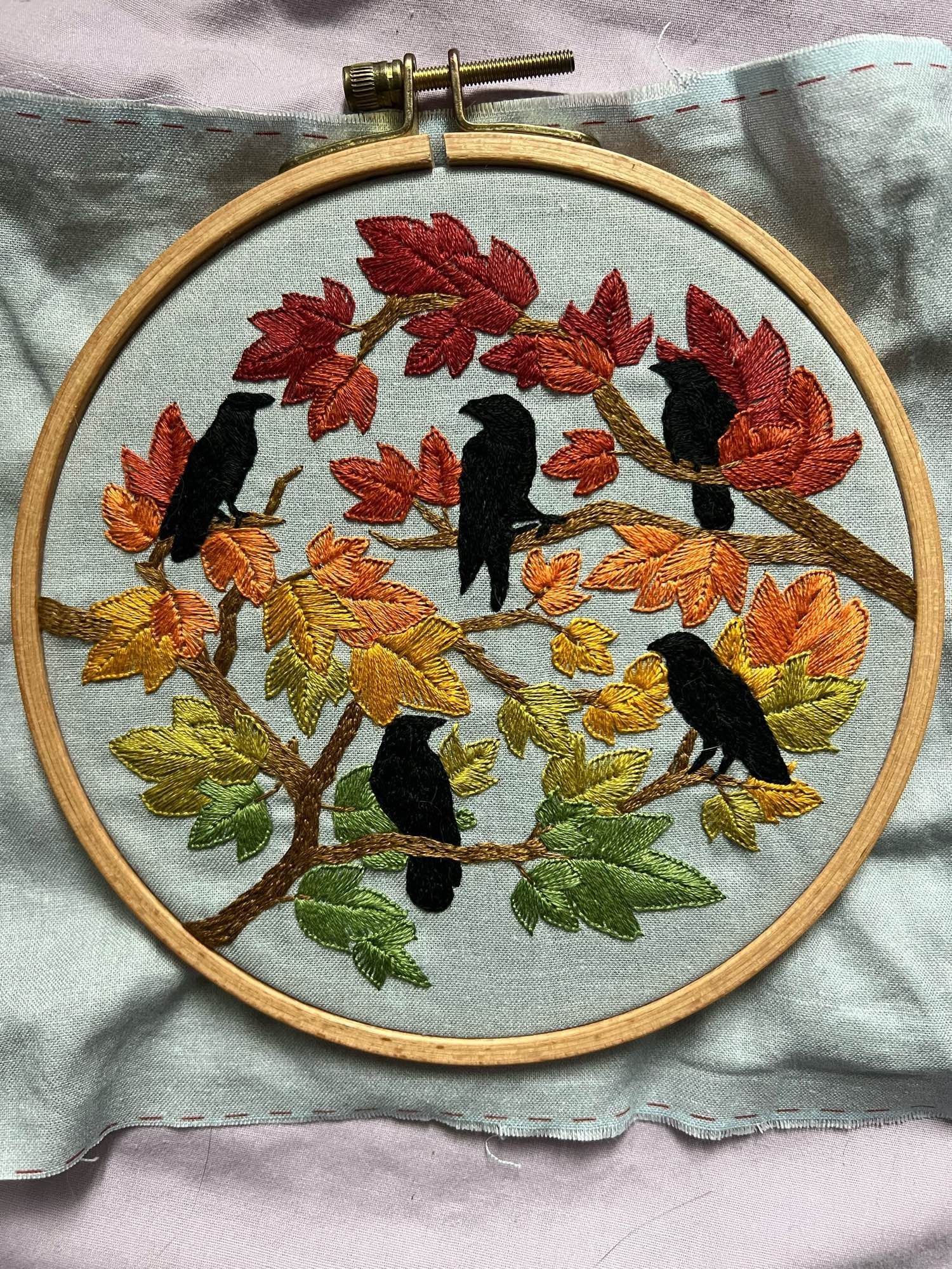 A 6 inch round embroidery hoop, showing a finished project: autumn birds by Jessica Long. Five featureless black corvids are perched in tree branches amidst a gradient of maple-style leaves. The leaves change gradient from green, to chartreuse, to mustard, to orange, to brick red, and finally deep red