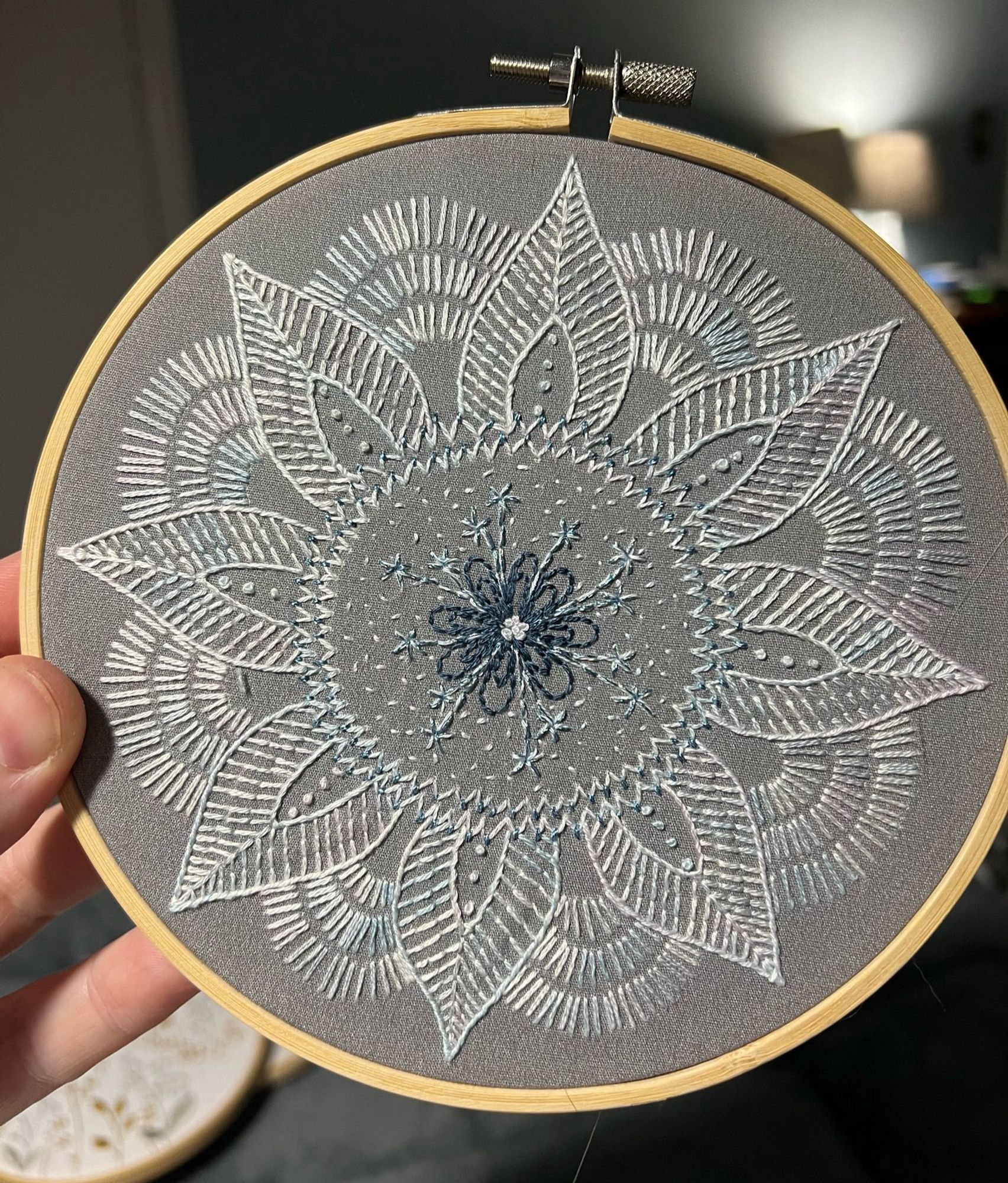 a 6” round flower-like mandala embroidered on gray fabric, primarily in a very pale variegated floss. designed by Cozy Blue at https://cozybluehandmade.com/collections/embroidery-kits