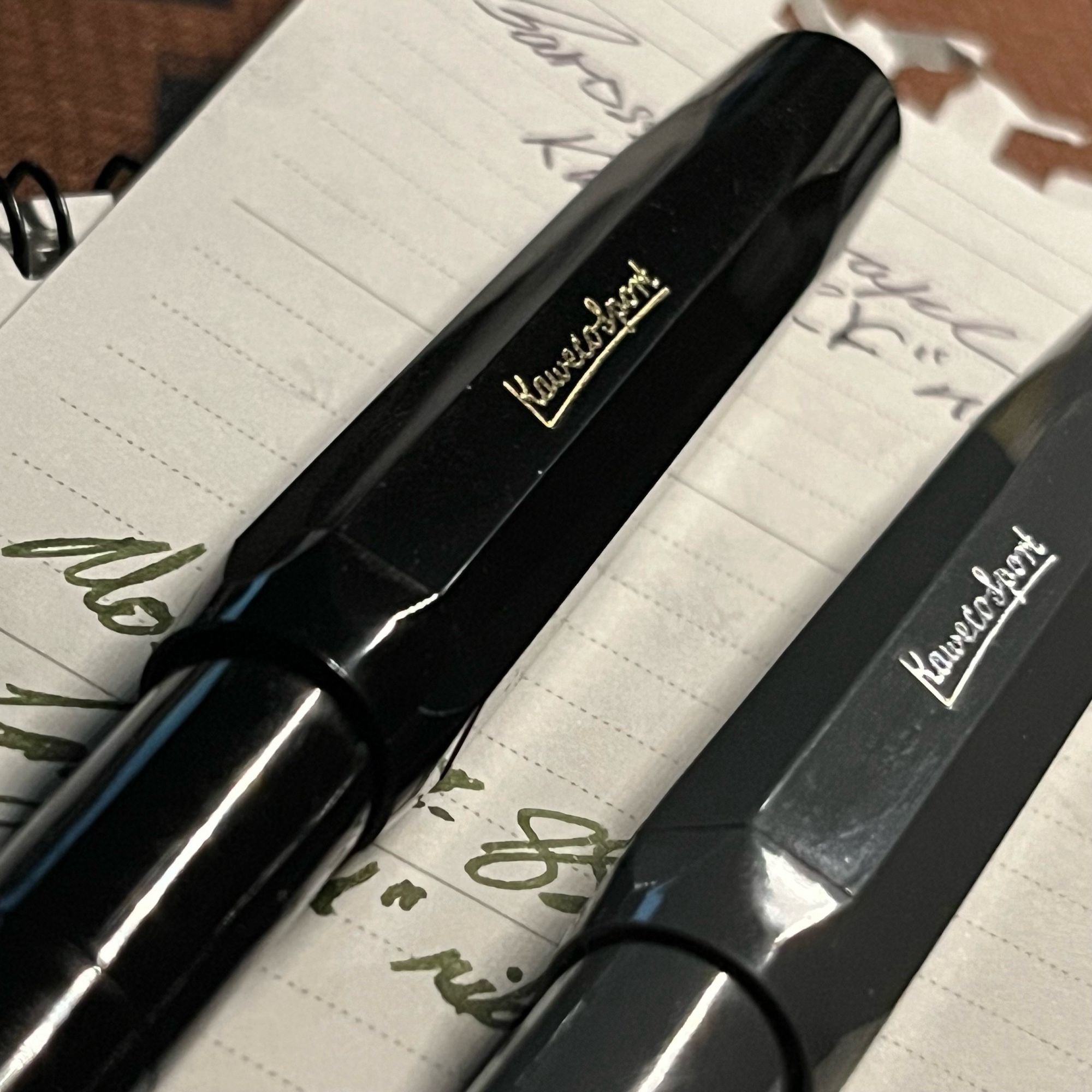 up close shot of 2 fountain pens. black kaweco classic sport EF inked w/ diamine’s writer’s blood near the middle and a grey kaweco skyline sport EF inked w/ robert oster’s barossa grape in the bottom right.