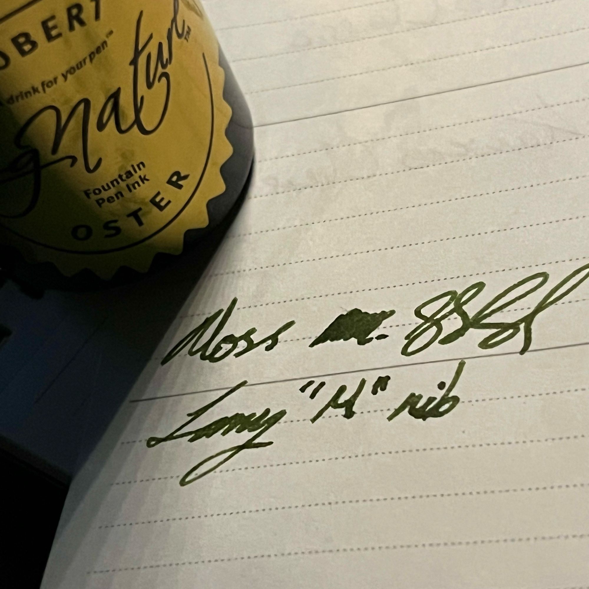 another corner shot of robert osters bottle in the top left corner that’s  sat on a writing sample of their moss green ink written w/ a lamy M nib near the middle.