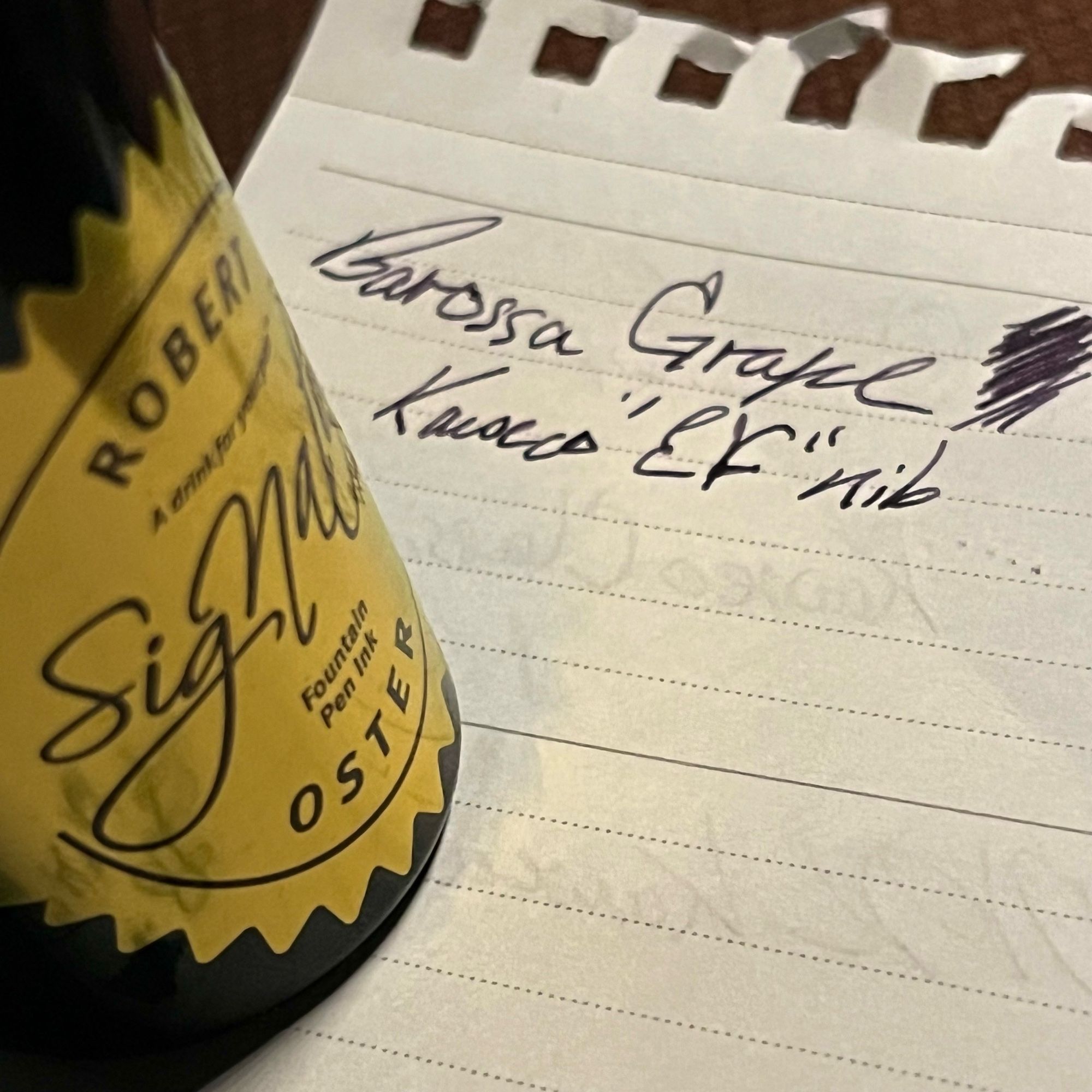 side shot of robert osters signature series brand labeled PET bottle on the left. sitting on a writing sample of their dark dusky purple barossa grape ink w/ a kaweco EF nib written on the upper half.