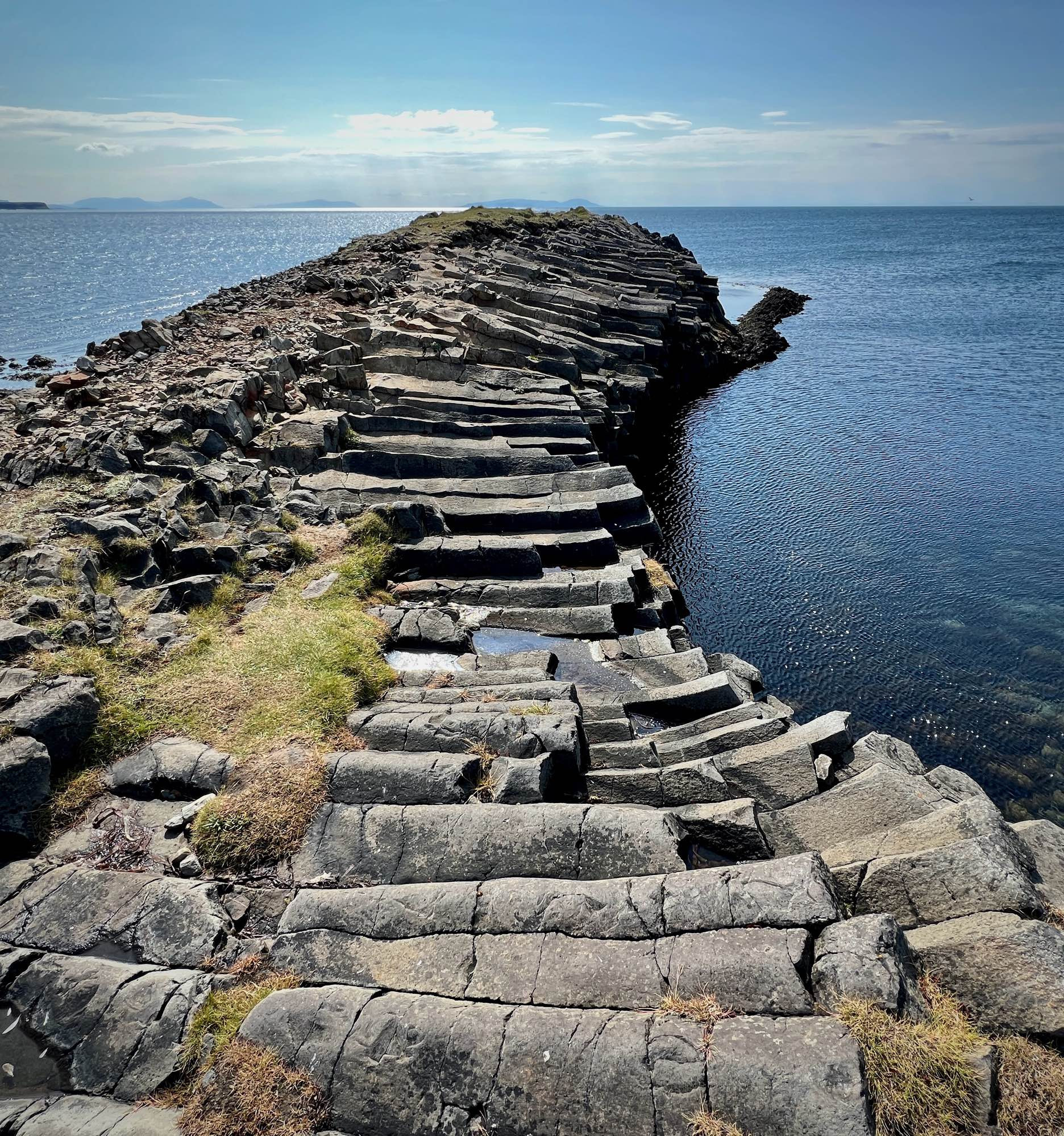 A small peninsula extends into the bay, its surface covered with basalt rock. On the right side, the land is entirely blanketed by horizontal basalt columns. These columns lie in front of you like the steps of a staircase, gently sloping downward at first and then rising again toward the end of the peninsula. 