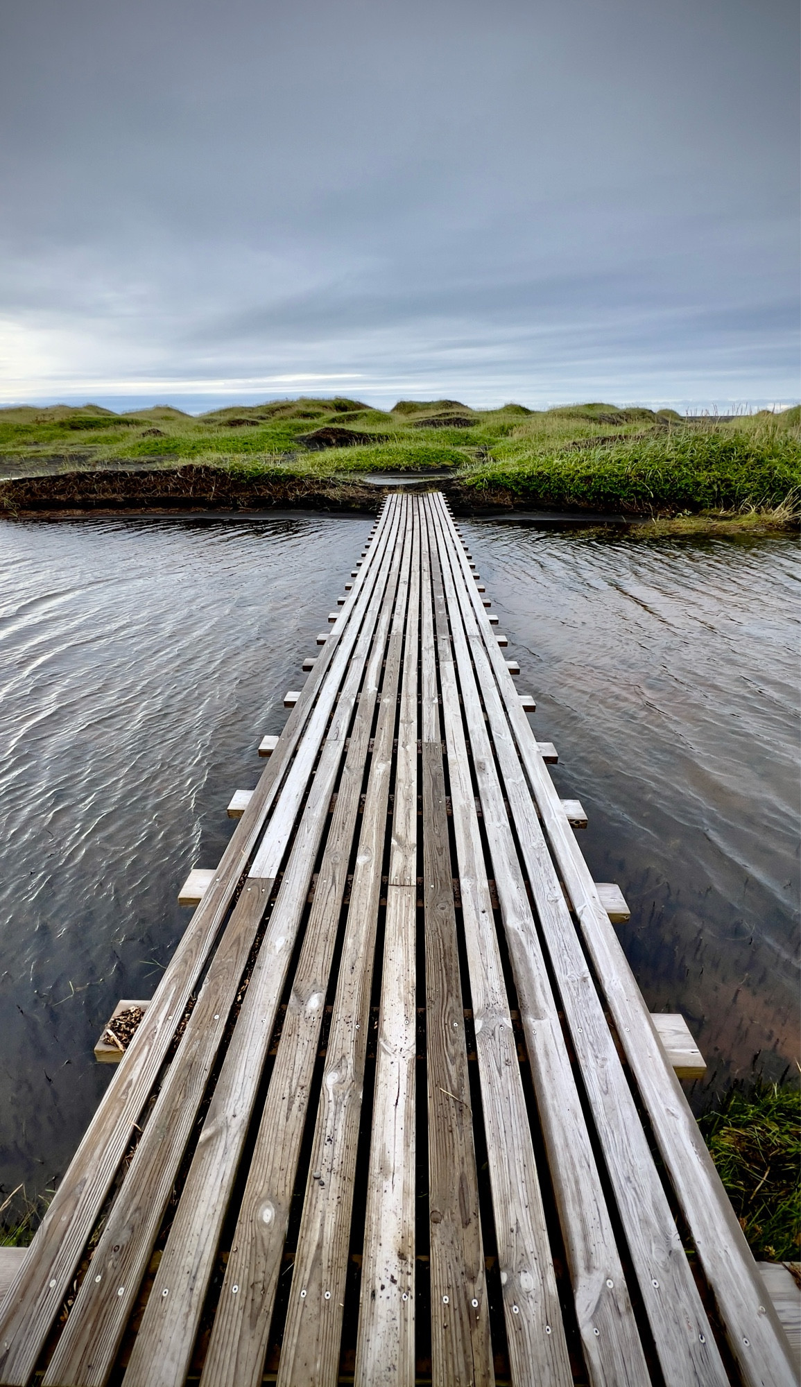 Imagine a long wooden footbridge extending across a small river. The planks of the bridge are laid lengthwise, creating a striped pattern that runs in the direction of the bridge’s length. On the opposite side of the river, the bridge leads into soft, green hills.