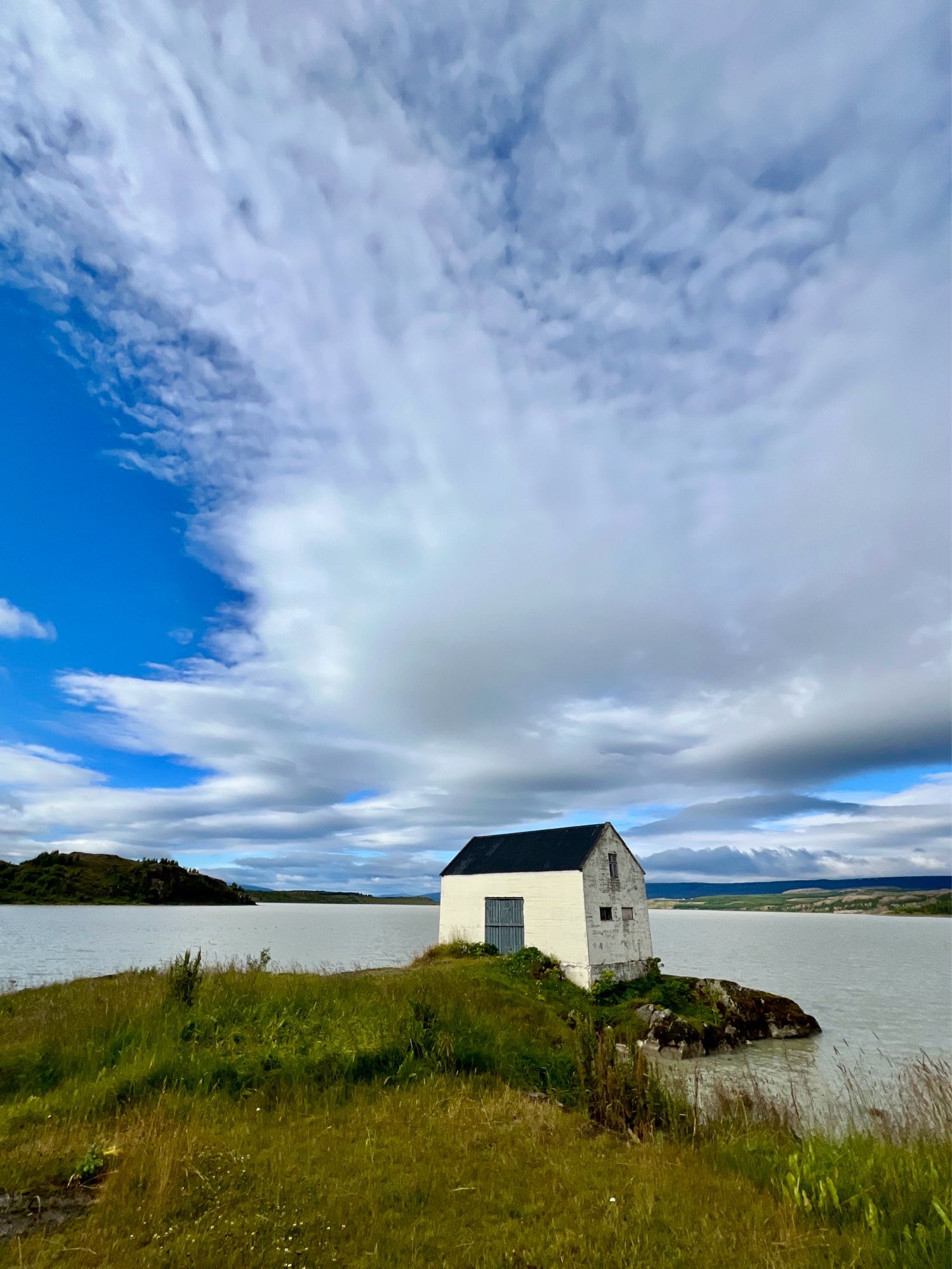 Imagine a cabin bathed in morning sunlight, standing on a narrow peninsula that stretches into a serene lakeshore. It is surrounded by vibrant green grass swaying gently in the breeze. You can hear the soft splashing of water against the shore. Overhead, the sky is a brilliant blue, with delicate, fluffy clouds drifting by, light and airy like strands of cotton candy.