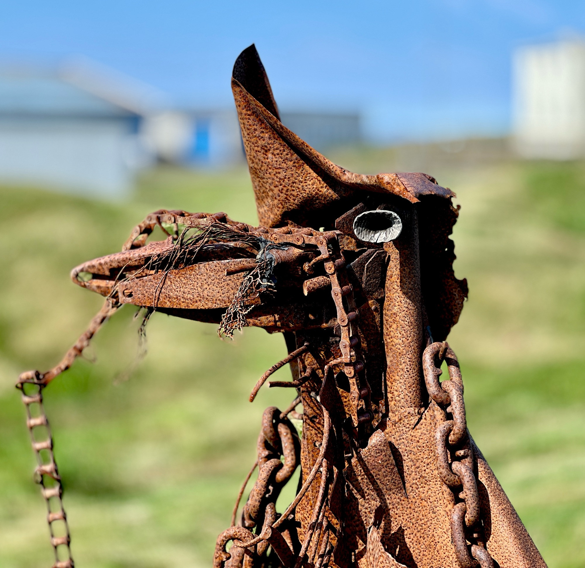 Eine abstrakte  menschengroße Skulptur, die einen stehenden Vogel mit langem Schnabel darstellen soll. Im Photo sind nur Kopf und Hals abgebildet. Die Figur besteht komplett aus verrostetem Metall, es sind auch schwere Gliederketten und eine Fahrradkette mit verarbeitet. Der verschwommene Hintergrund zeigt eine saftig grüne Wiese mit Häusern am Rande. 