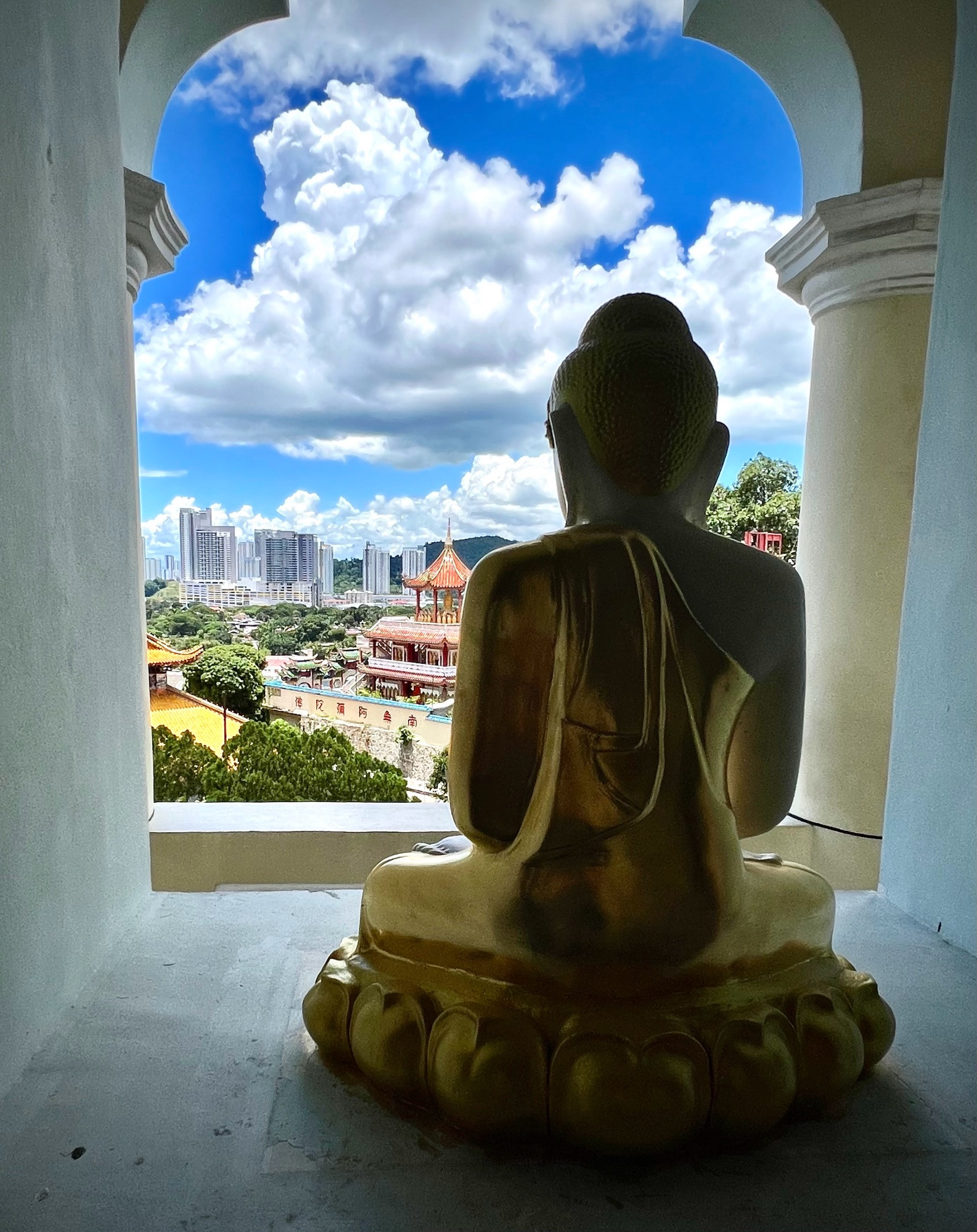 Imagine a golden Buddha statue seated in an opening within a white stone wall. You see the back of the statue as the Buddha faces forward, looking out onto an ancient temple complex in the foreground. Beyond the temples, you can see tall, modern skyscrapers rising in the background. The contrast between the traditional, spiritual setting of the temple and the towering, modern buildings represents the juxtaposition of the old and the new, the sacred and the urban. The Buddha sits calmly, as if observing the passage of time and change.