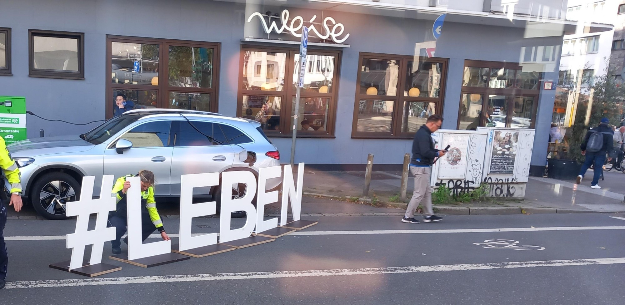 Pappaufsteller als Schriftzug #LEBEN auf einem Radweg, ein Polizist stellt ihn gerade auf. 