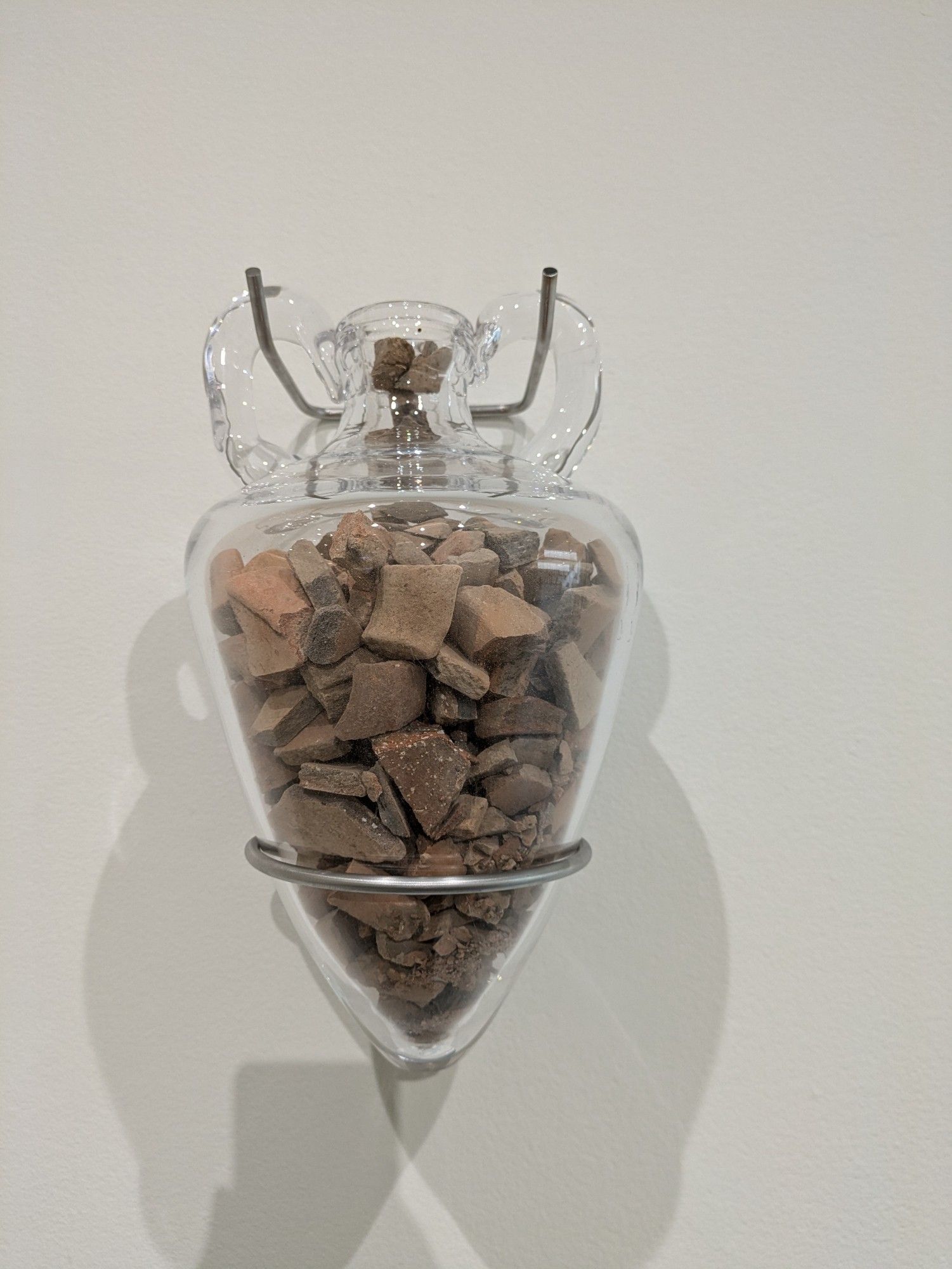 a glass amphora form suspended by the handles from a forked metal wall mount. it is filled with shards from 10th-c. pottery.

Saj Issa, If I Don't Take It Someone Else Will, 2024