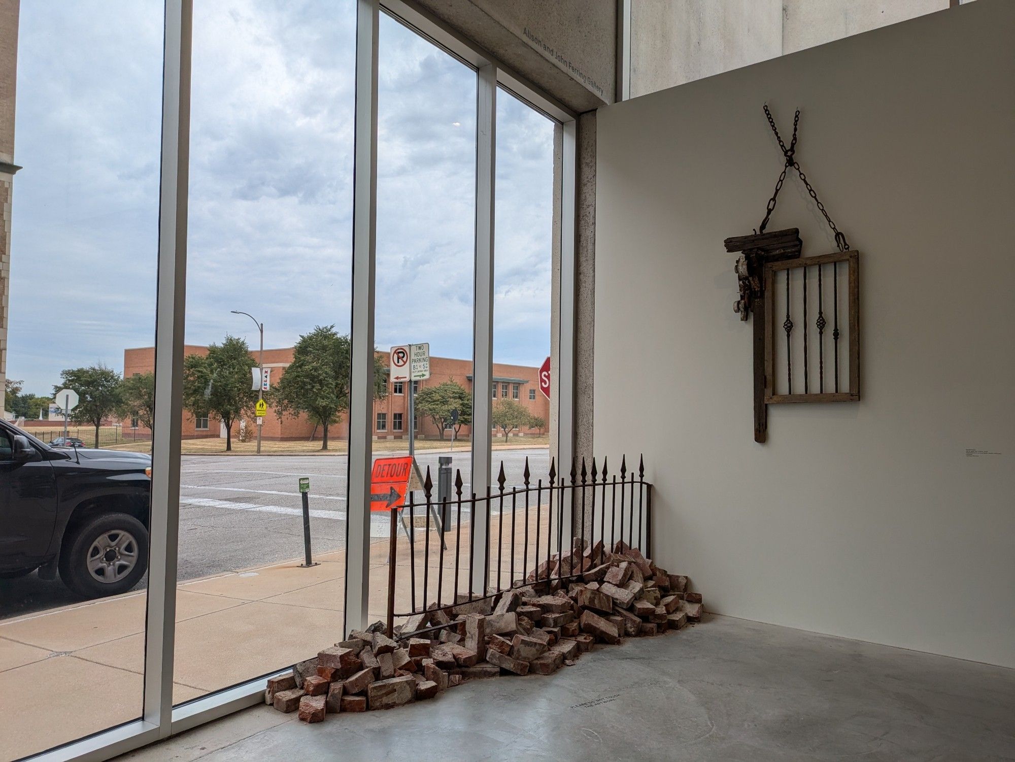 a corner of an airy boxy room with one wall made of glass and the other a white wall, slightly shadowed on an overcast day. on the wall a wood and iron casement with a partial external frame hangs suspended by slavery-recalling chains. in the corner, bricks from the underserved, burned neighborhoods nearby (in the direction of the corner, actually) lie in a broad, haphazard pile along a crooked fragment of iron fencing with pointed finials (Ronald Young, Out of Sight--Out of Mind, 2018)