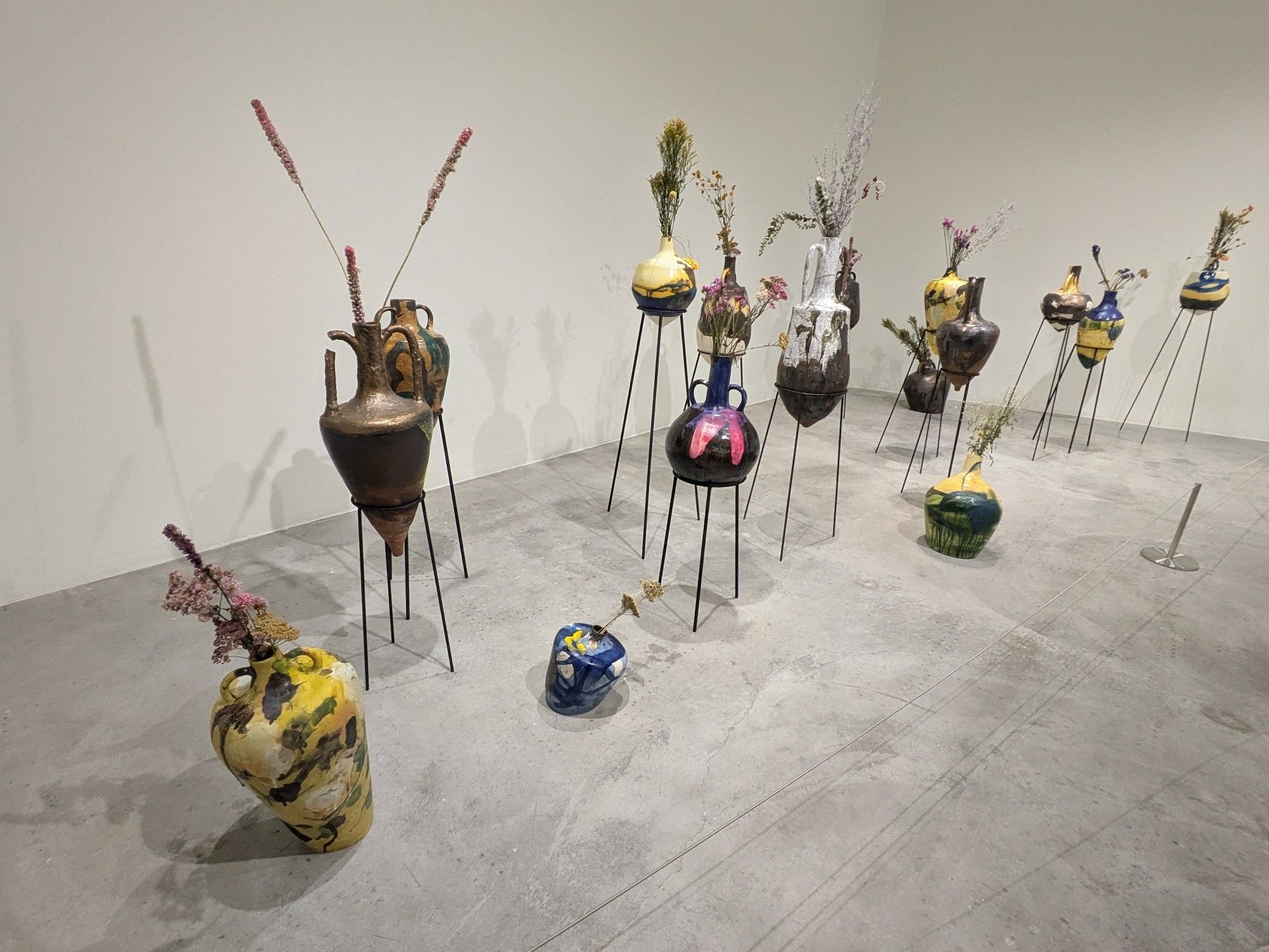 many amphorae painted in various colors including bright yellow swathes and filled with long, tall flowers in a white room with a concrete floor. most of the vessels are mounted on tripods but three, two noticeably misshapen, sit directly on the ground.

Saj Issa, Landscape Amphorae, 2024