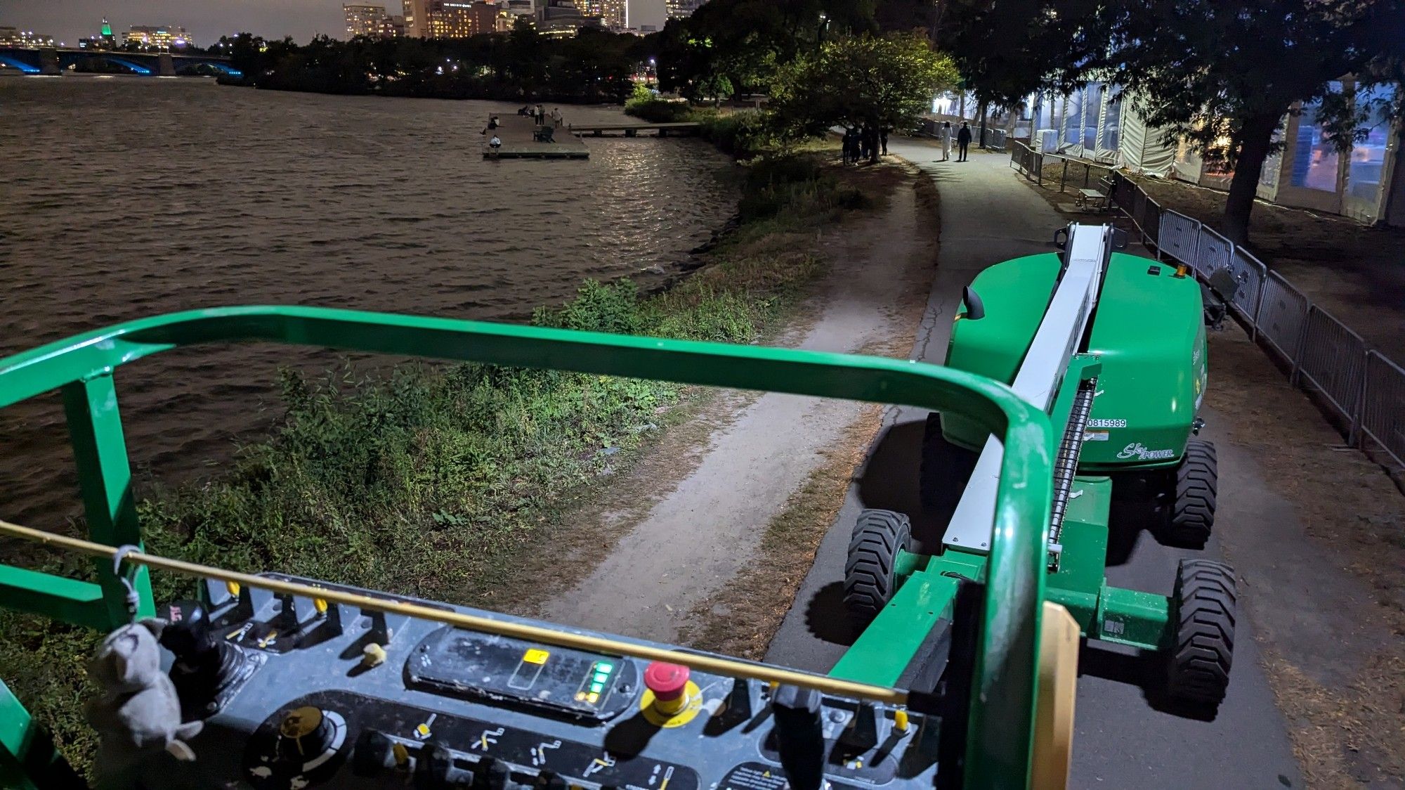 A point-of-view from the bucket of a boom lift being driven along a path about ten feet from the river's edge. In the background, the city skyline can be seen lit up at dusk, and along the river a large event tent with crowd control barriers is set up.