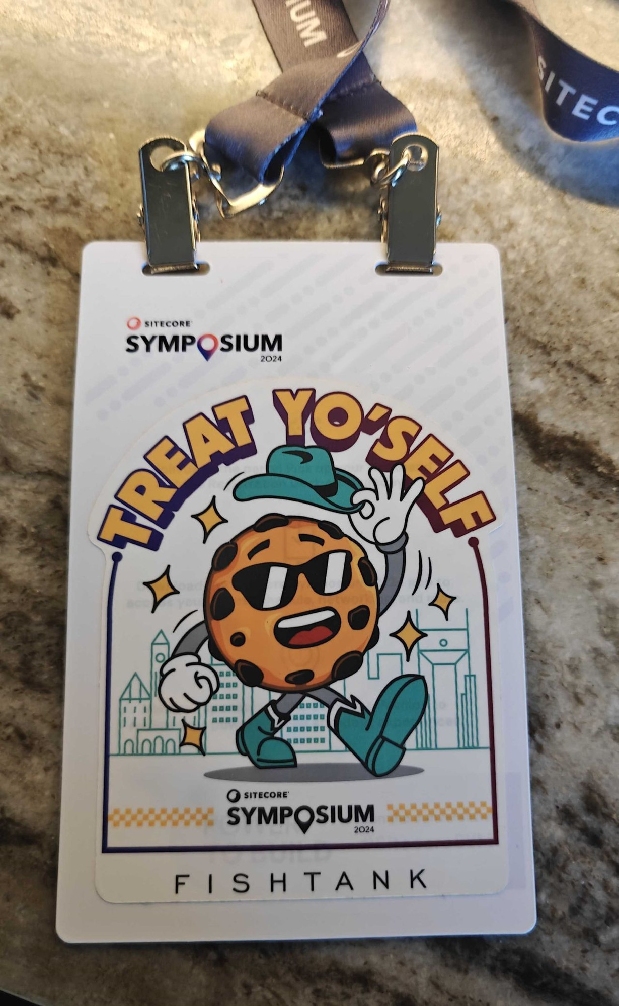 A lanyard for Sitecore Sympsium lies upon a marble tabletop. A picture has been placed overtop the lanyard that features a cookie dressed in cowboy boots and tipping their cowboy hat. The text above the cookie reads "Treat Yo'Self".

The label at the bottom of the sticker reads "Fishtank".