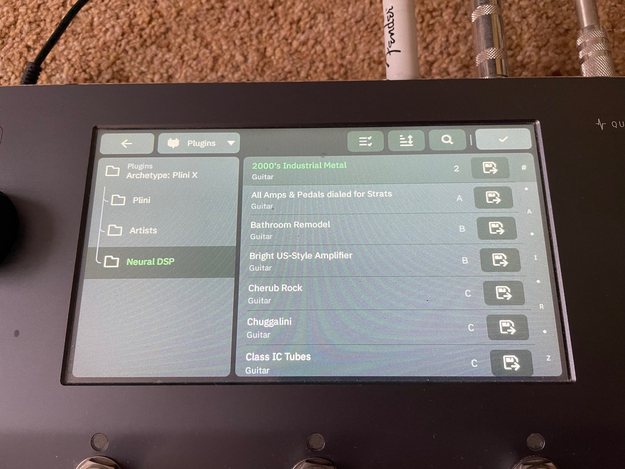 A NeuralDSP Quad Cortex sat on the floor, powered up, showing a closeup of the touchscreen display. The “2000’s Industrial Metal” preset has been selected from the “Archetype: Plini X” plugin, under the “Neural DSP” folder.