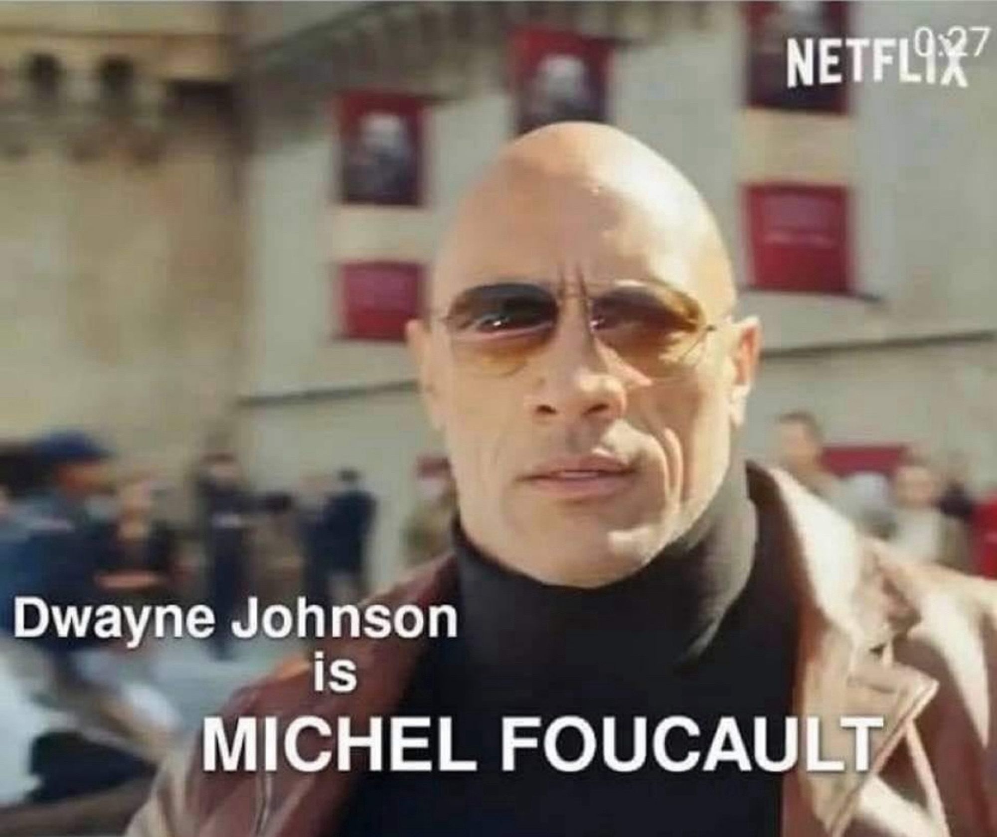 The rock in sunglasses, a black turtle neck, and leather jacket in front of a blurry crowd in a public square