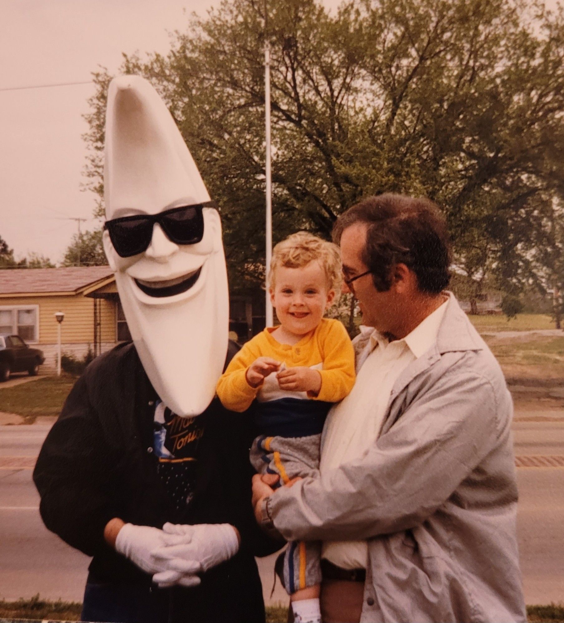 A person dressed as McDonald's ad icon, Mac Tonight, with a moon shaped head and sunglasses. Next to him is a dad with dark hair holding a blond curly haired toddler boy grinning at the camera.