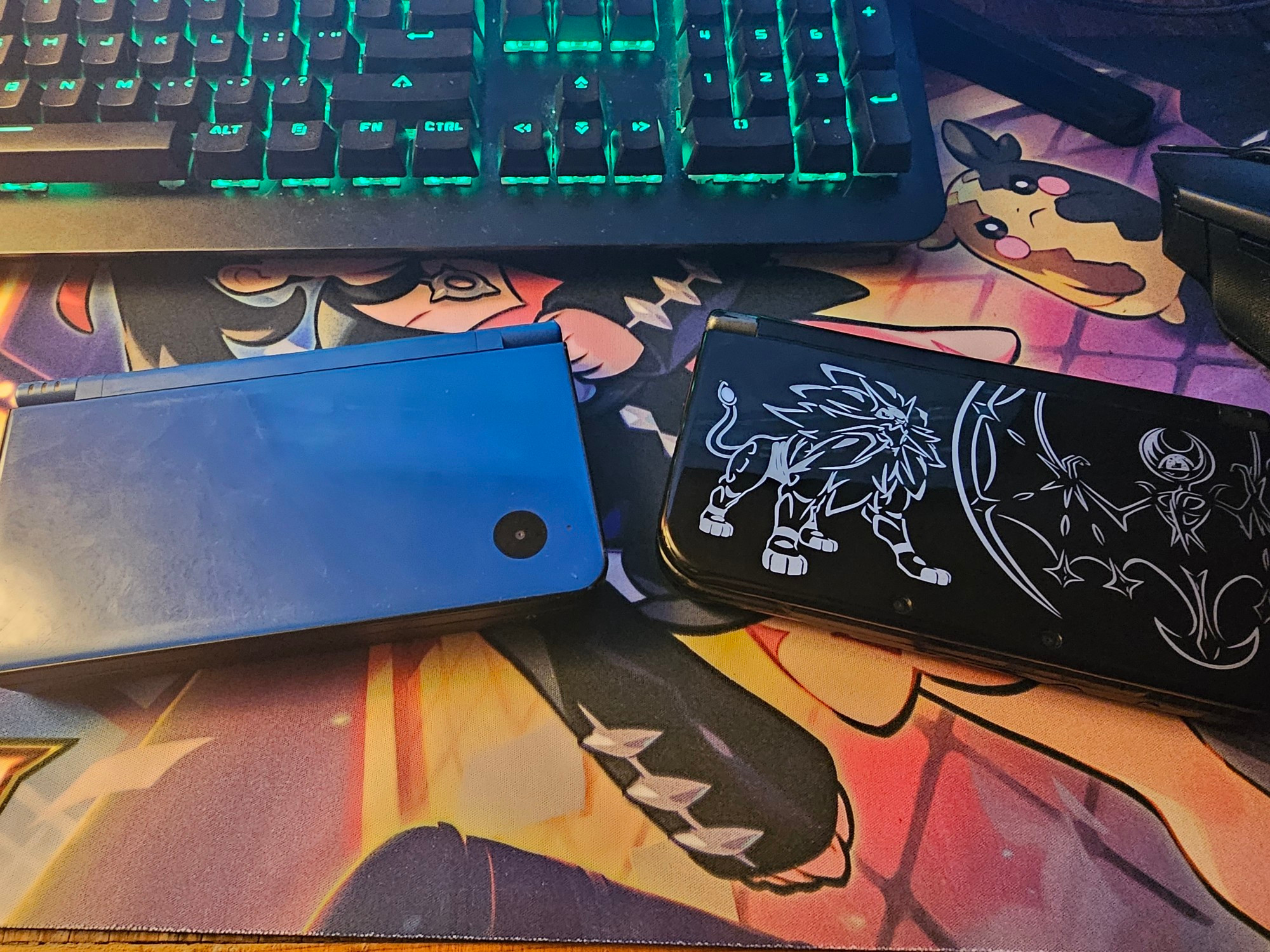 A blue DSi XL and black New 3DS XL with Solgaleo and Lunala on it in white, both sitting on a TCG playmat of Marnie from Pokémon Sword and Shield, but her face is being covered by an RGB keyboard, Morpeko however is in full view