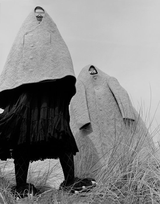 Andrew Nuding, 2019, from series "Making Strange" — black and white photo of 2 figures outdoors standing amid grasses. In the foreground, wearing a short, oversized felt cape with no arms with small opening for face (wearing sunglasses), a black voluminous calf-length skirt, black tights and bear slippers with 3 claws each. In the background, a much longer, ankle-length, oversized cape with arms (but arms are not in it) and same face-hole (and sunglasses).