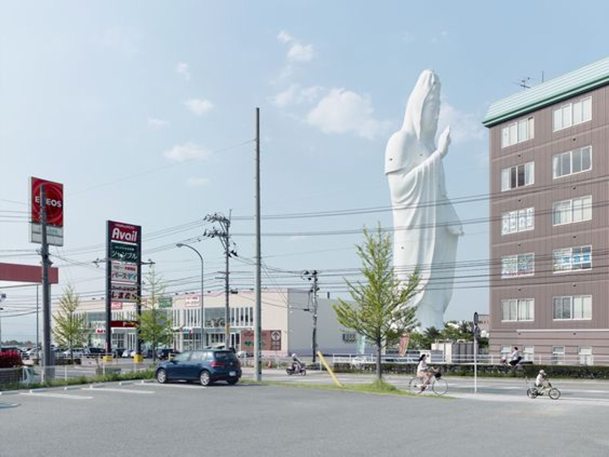 Fabrice Fouillet, Daikannon, 2019, Sendai, Japan (330 ft) — stunning, white, monumental figure against a pale blue sky with a few clouds, set behind an everyday setting of buildings, a fuel station, light poles, road, cars, people on bicycles. The bodhisattva Kannon (known in Chinese as Guanyin and Avalokitesvara in Sanskrit) is the bodhisattva associated with compassion.