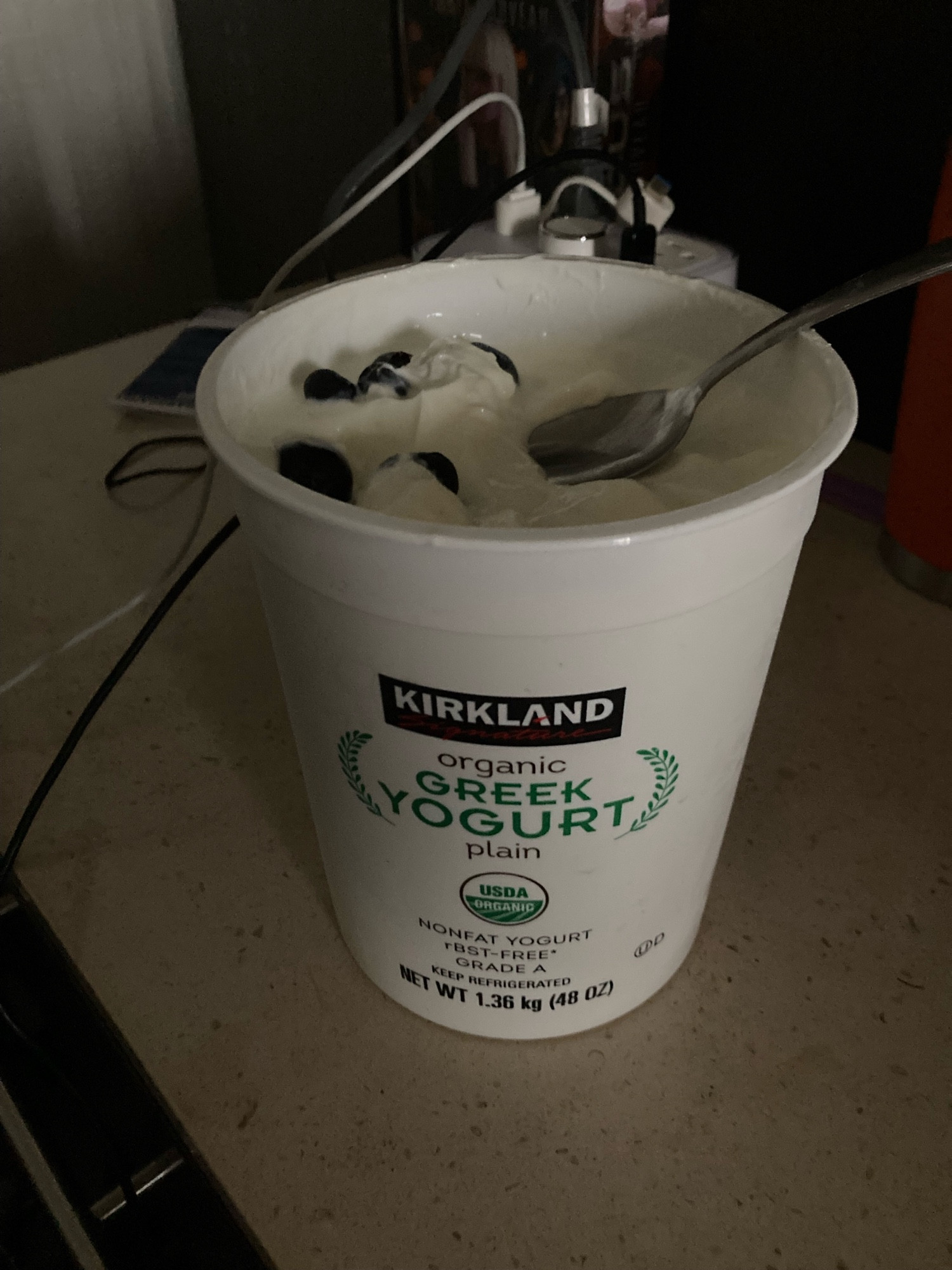 A 48 oz tub of Greek yogurt with a handful of blueberries on top and a spoon.