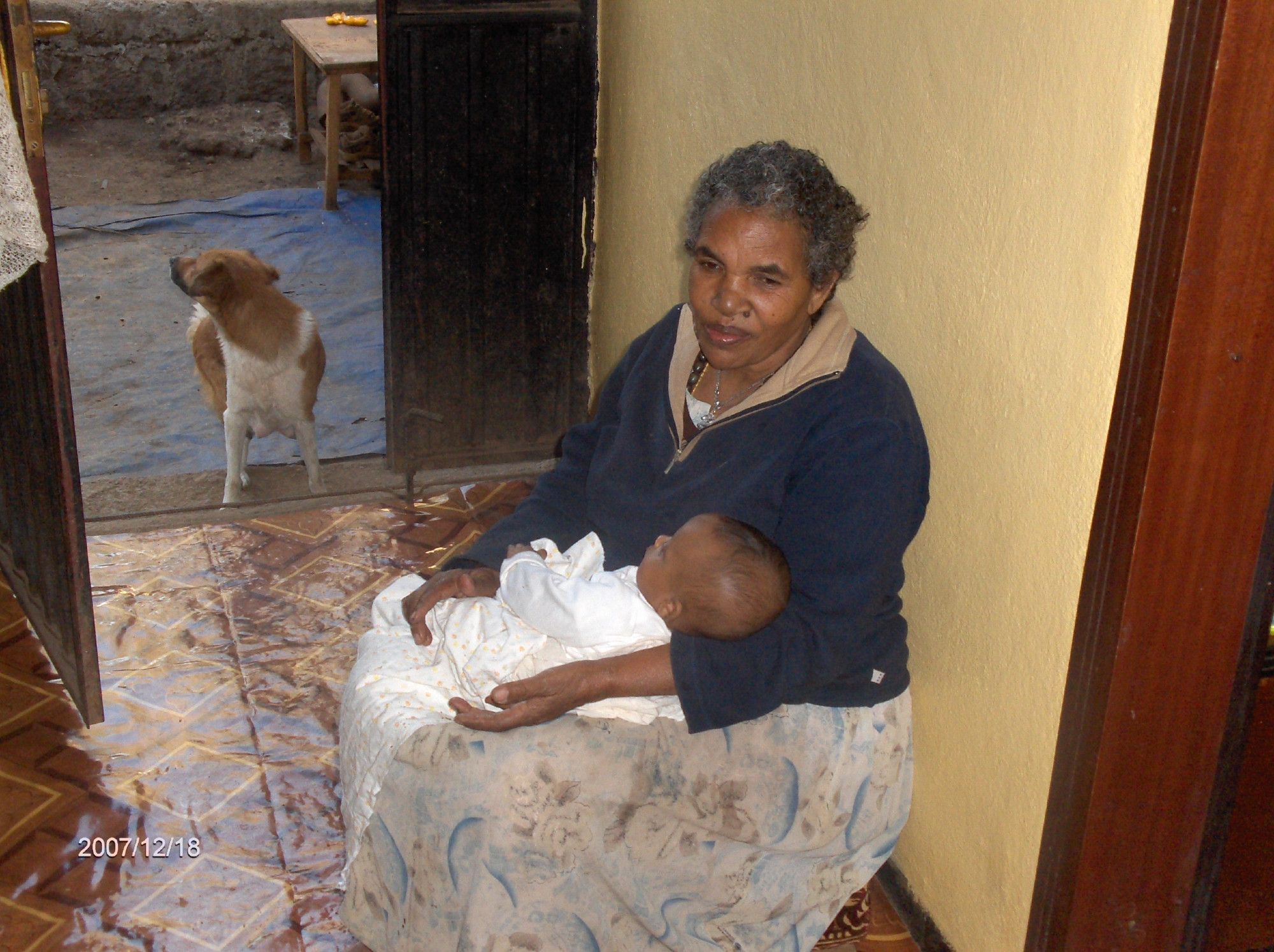 The same lady sitting inside the house with a little grandchild on her knee