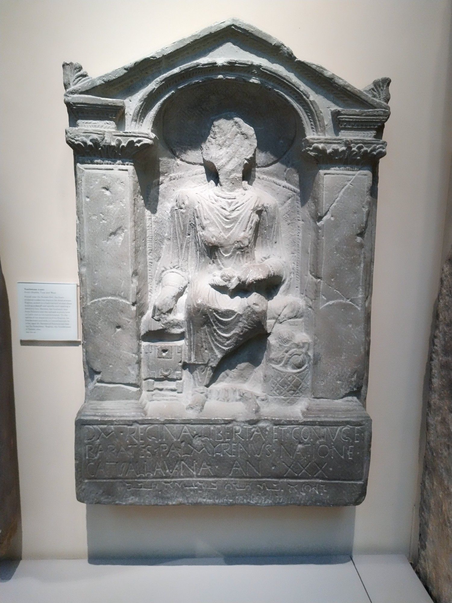 A tombstone showing a woman seated, in an apse. Her face is missing.