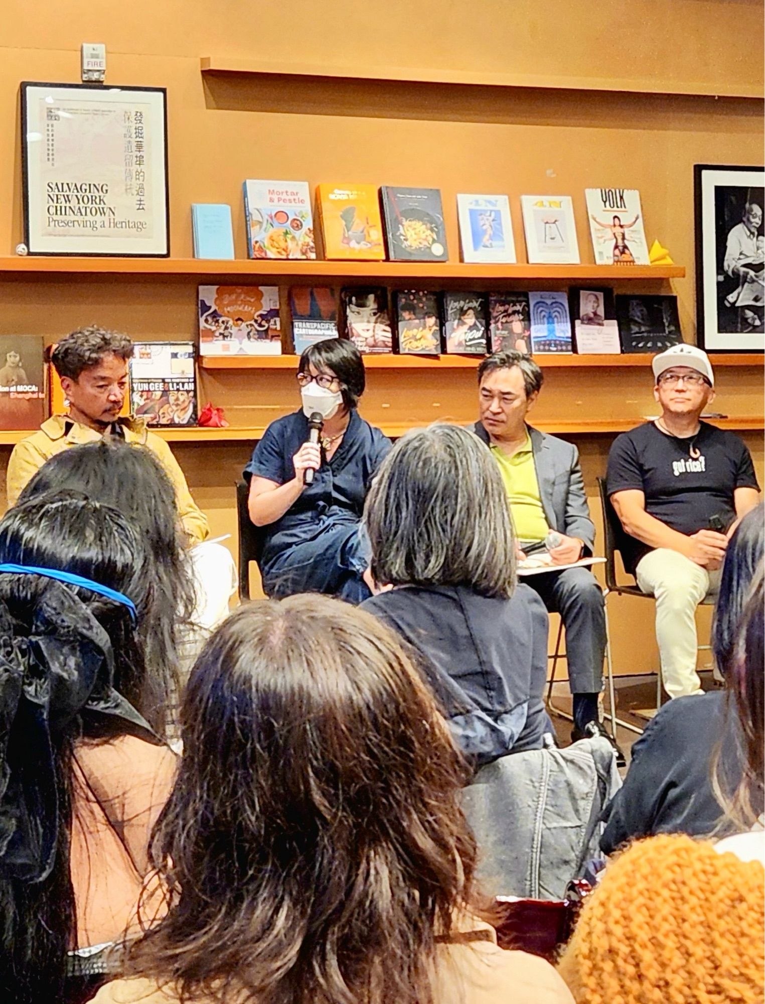 Melissa, wearing a mask, speaks into a mic next to her fellow panelists, 3 men, in front of an audience. Everyone is seated.