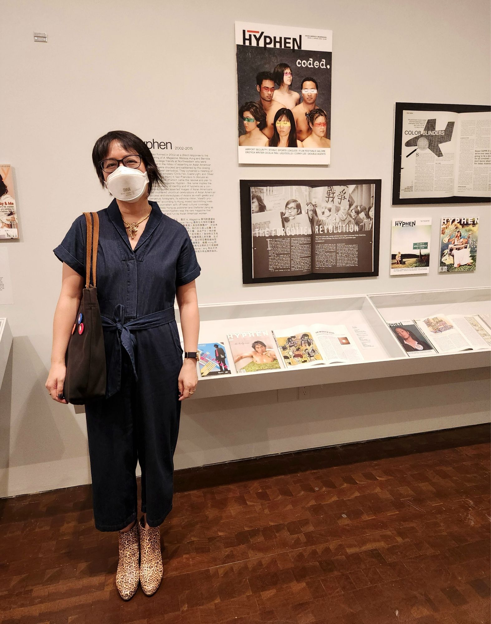 Melissa, wearing a mask and a denim jumpsuit, in front of the Hyphen section of an exhibit in a museum gallery. There is an enlarged cover of an issue of Hyphen on the wall.