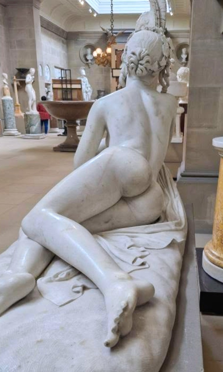 A statue of a recumbent reveller, having a little rest in between revels, in a grand room with lots of other sculptures 