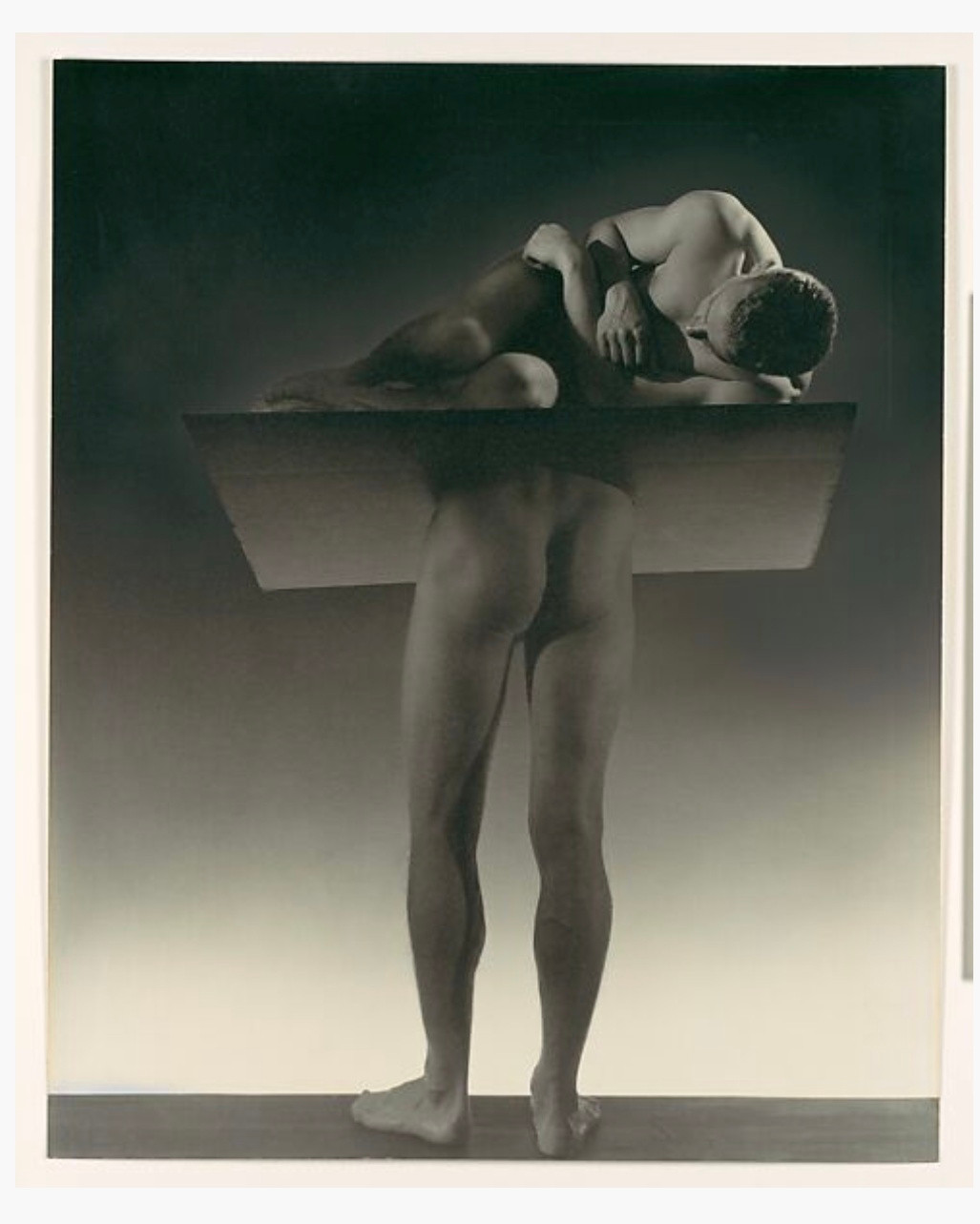 An abstract photo showing a figure curled in the foetal position on top of a platform that intersects the bottom half of another figure