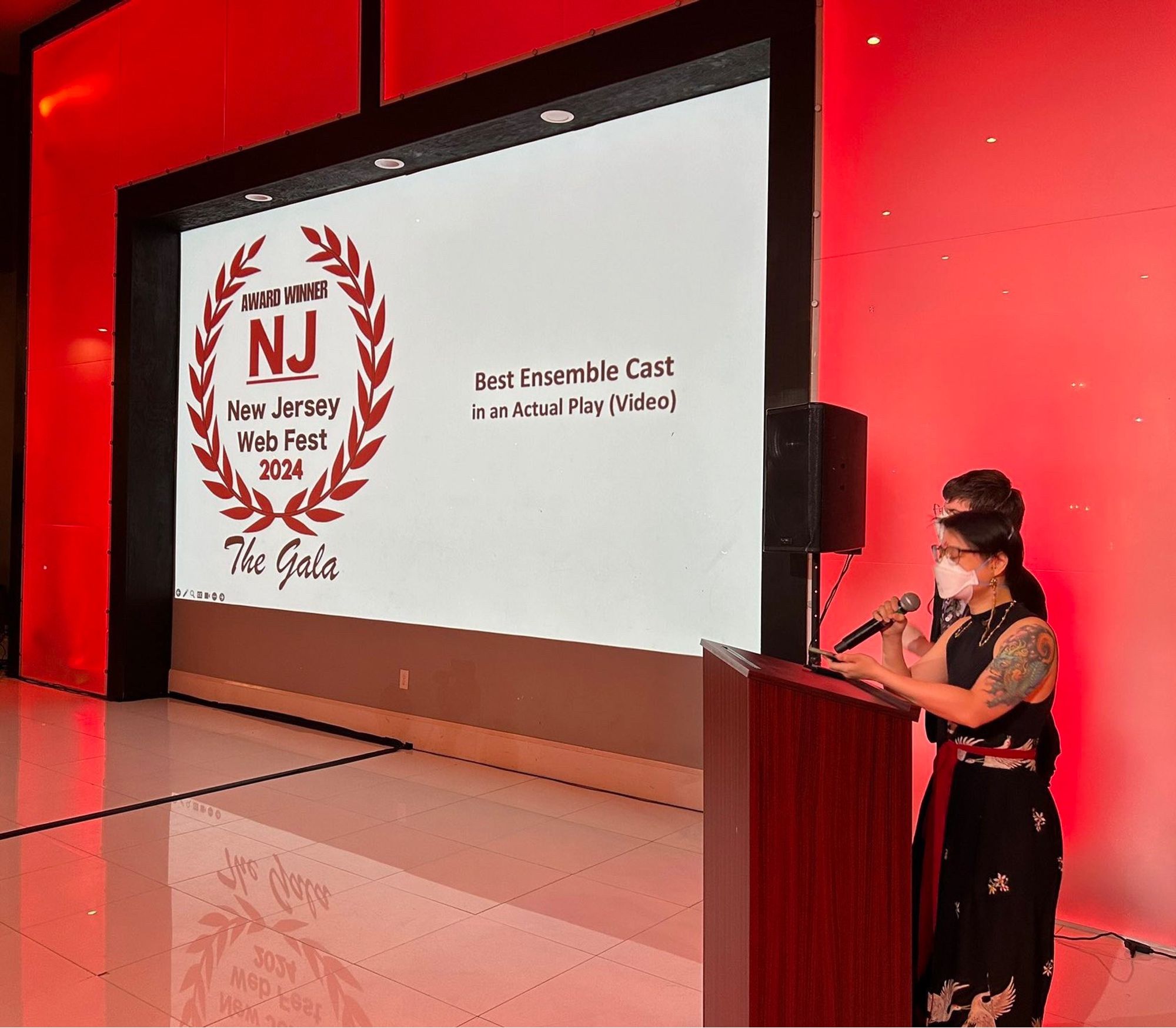 A photo of Connie and Sea at the NJ Webfest Awards Gala. The photo features Connie and Sea standing behind a podium with a microphone. On the left is a large screen projecting the NJ Webfest Logo and text that reads: “Best Ensemble Cast in an Actual Play (Video).” There is warm red lighting across the photo. Connie is dressed in a hanfu skirt and black sleeveless top, and though he’s covering most of Sea’s body, they can both be seen wearing white N95 masks.