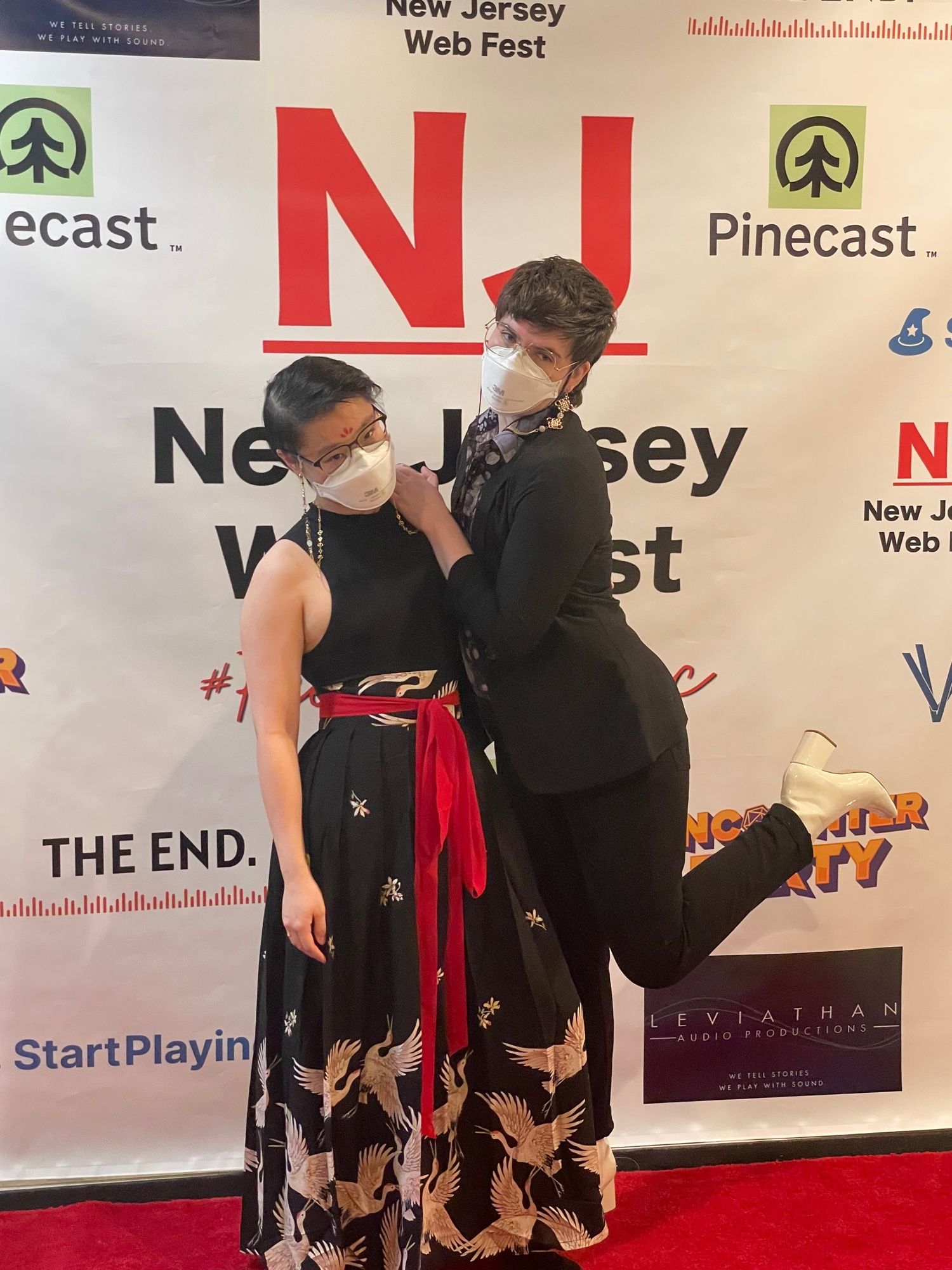 Connie (left) and Sea (right) pose in front of the New Jersey WebFest red carpet. connie wears a sleeveless black tank top and a black hanfu skirt with gold cranes and a red sash, as well as a golden glasses chain. Sea wears a black jacket over a black and purple button-up shirt and black slacks, as well as white platform shoes. Sea leans on Connie with their leg kicked up. They are both wearing white N95s.