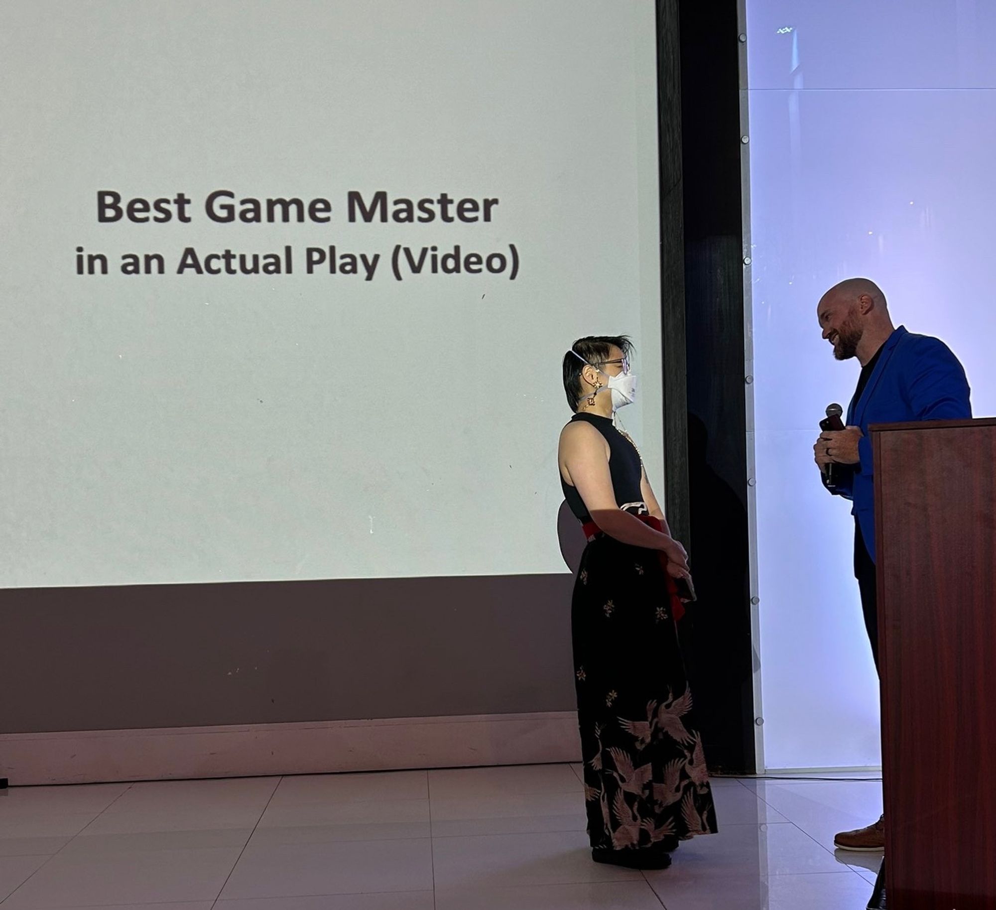 Connie stands in front of a large projection screen that reads, “Best Game Master in an Actual Play (Video).” They are facing Ned Donovan, who stands behind a wooden podium. Connie has short black hair shaved on one side and wears an N95, a sleeveless black tank top, and a black hanfu skirt with golden cranes and a red sash. Ned has a shaved head and a beard and wears a blue dress shirt, holding a microphone while smiling at Connie.