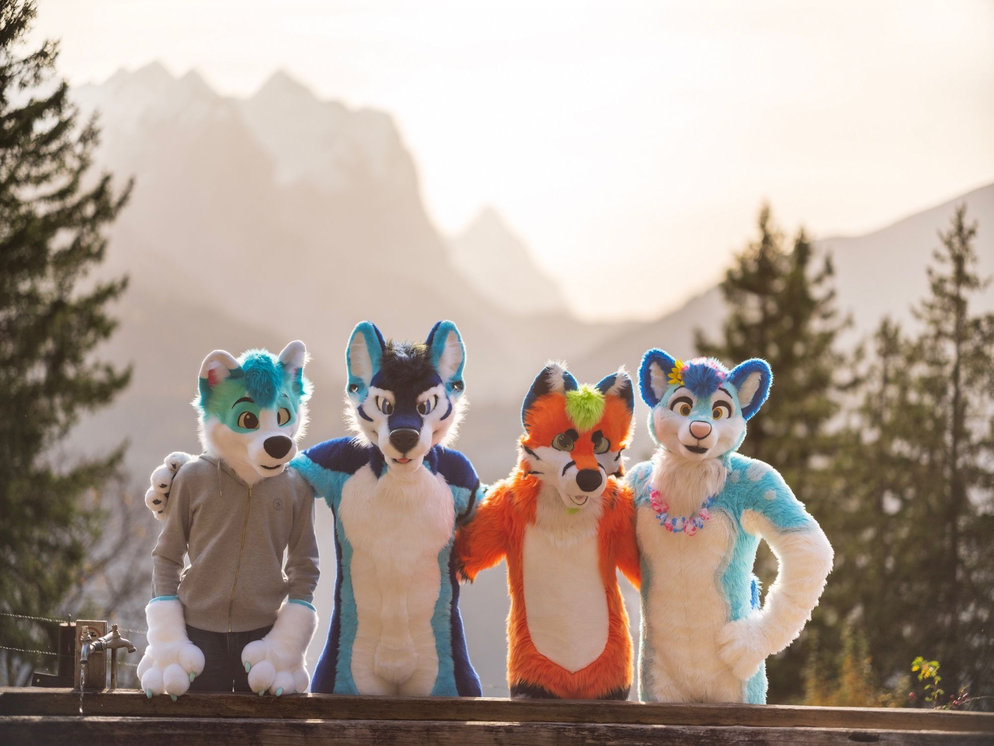 Four fursuiters next to each other in front of a wooden drinking trough, with trees and a faint mountainside in the background.