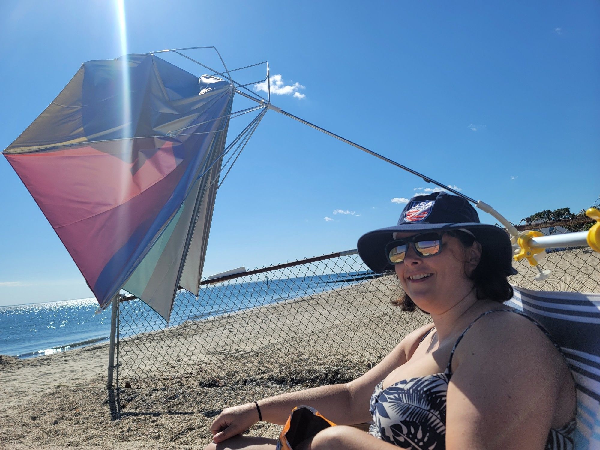 A photo on the beach of the op (a tubby white lady) smiling in a bathing suit and sun hat, as a chair-mounted umbrella has been turned inside out and badly damaged by the wind