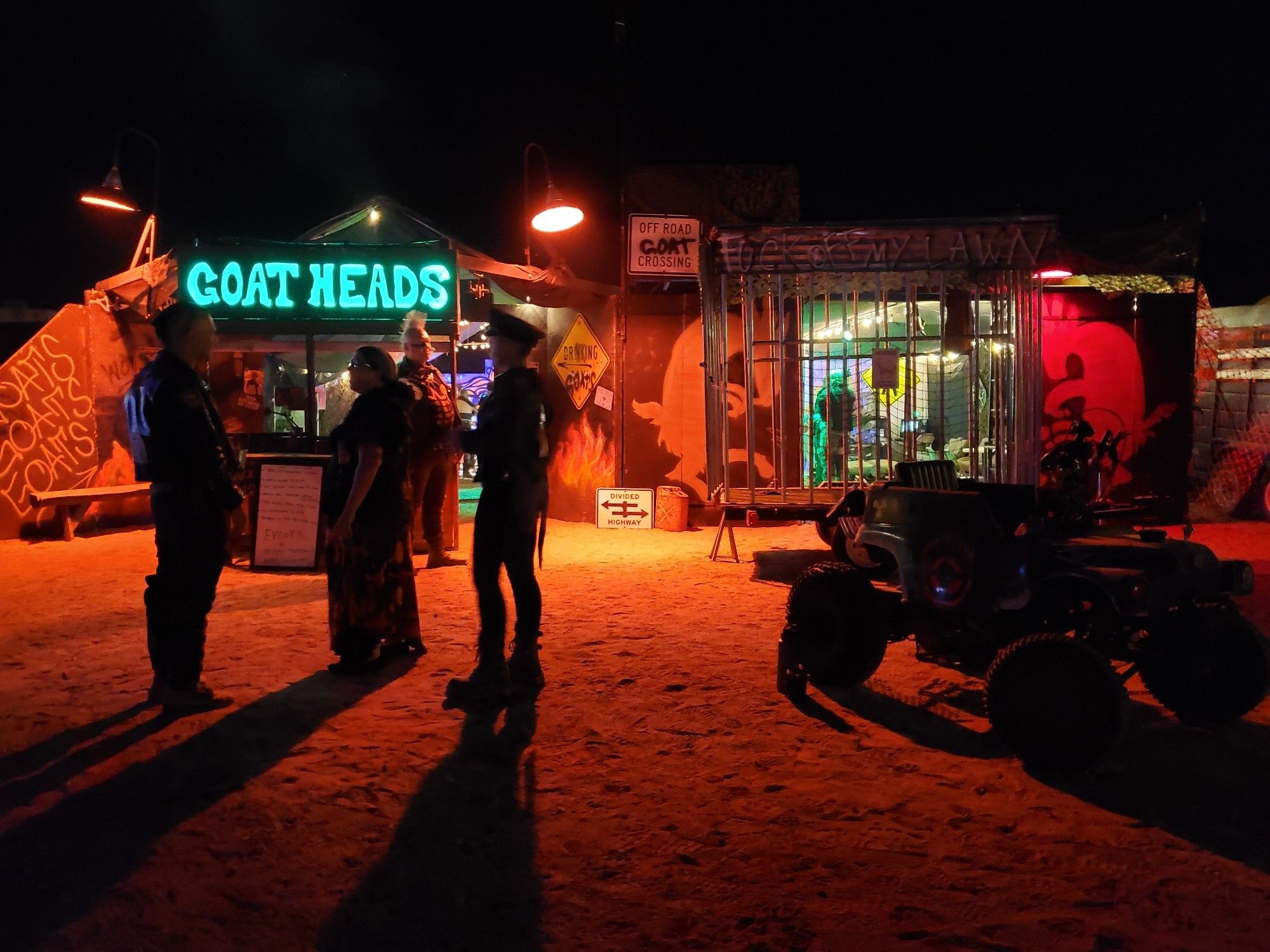 night scene with intense red light cast on desert sand. people stand around in leather and weathered cloth, wasteland attire. a souped up buggy is in the foreground and a cage and a neon green sign with "goat heads" emblazoned on it in the background.