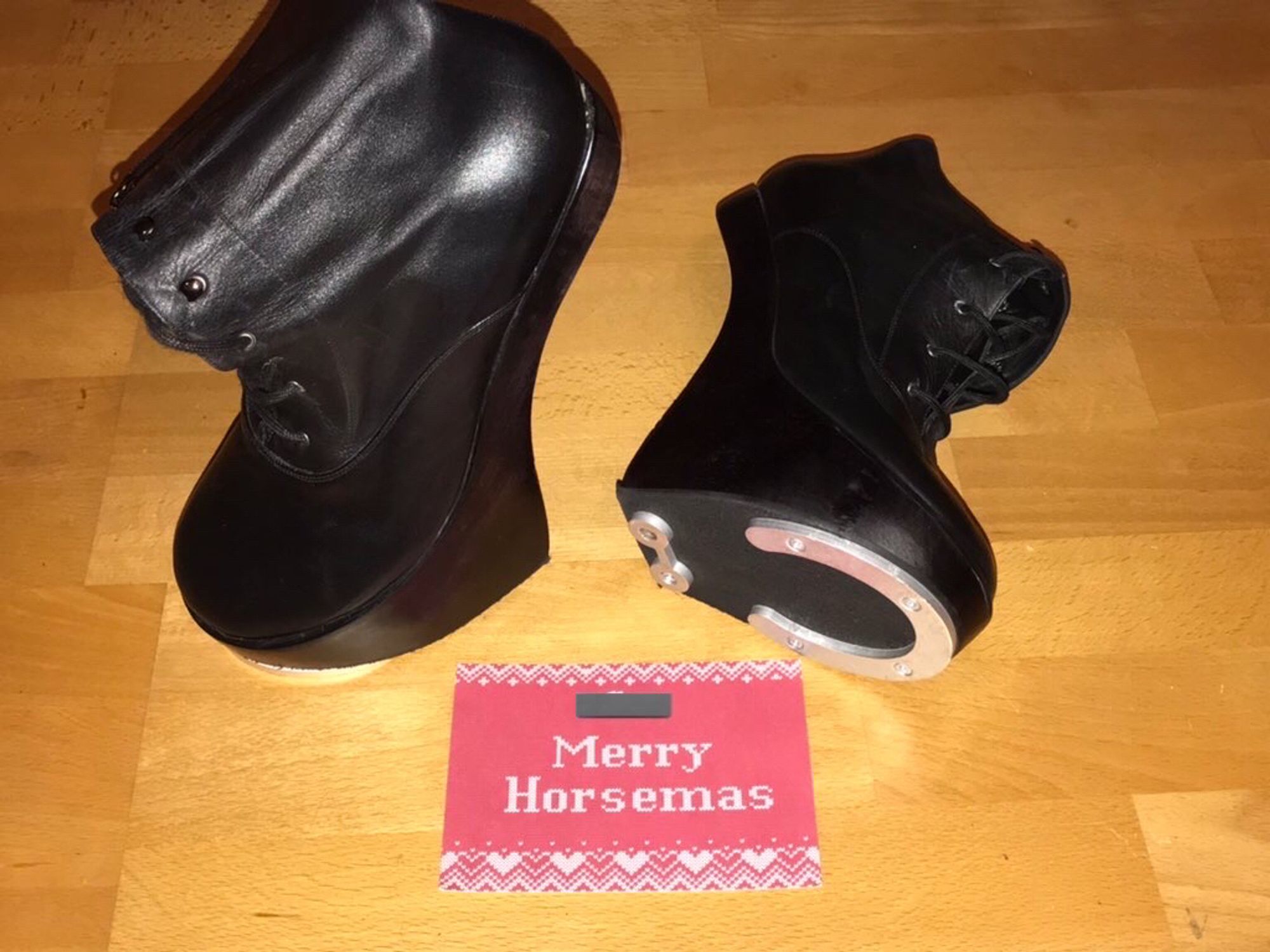 A postcard which says “Merry Horsemas” in a white font on a red background, with some white patterning top and bottom.  (The print resembles stitches in a Christmas jumper.) A pair of black leather pony hoof boots is placed next to the card; one boot is lying down and showing off the metal horseshoe screwed onto the wooden sole.