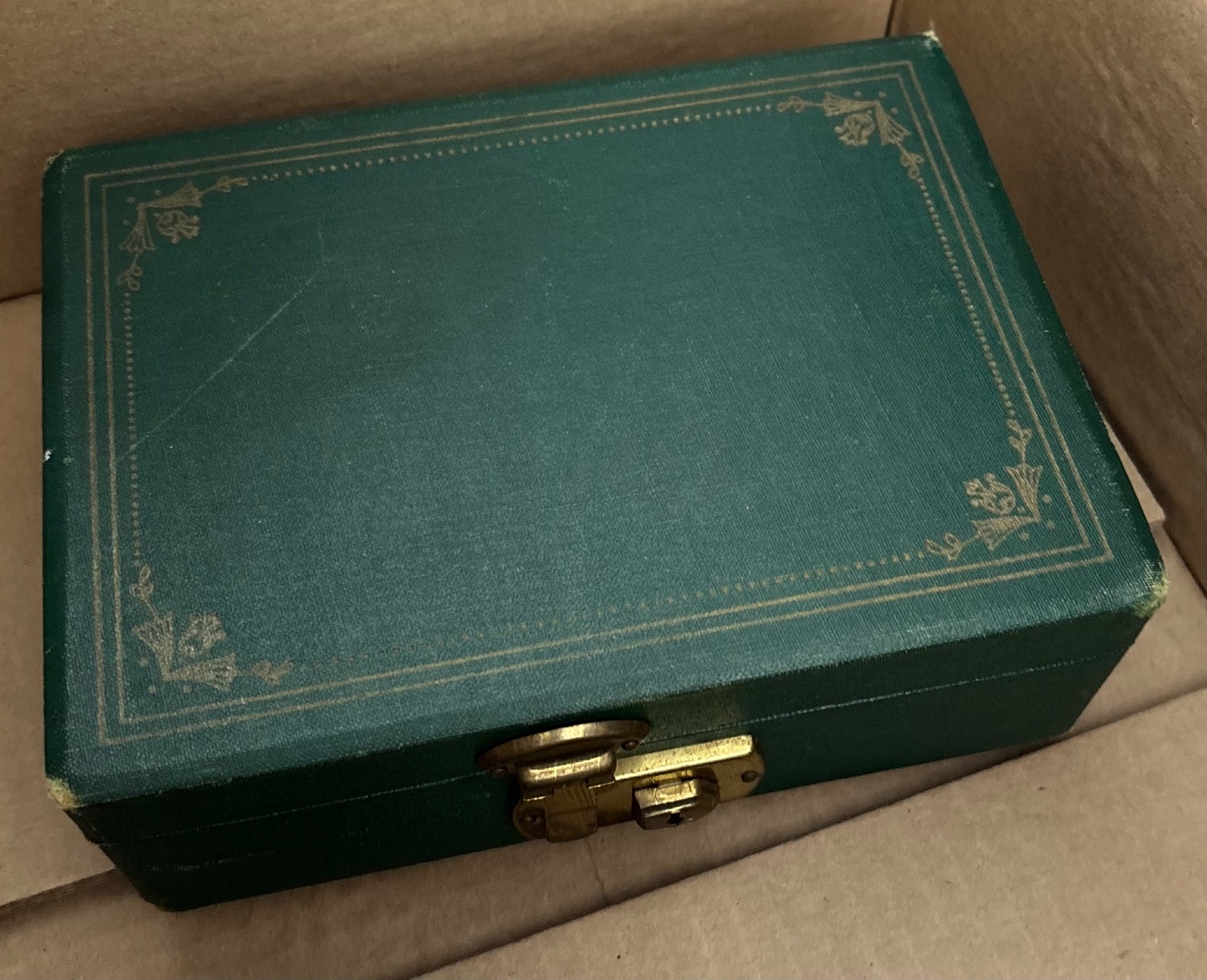 Photo of a dark green jewelry box inside a cardboard box. The latch is closed, and made of cheap (but gold-colored) metal.